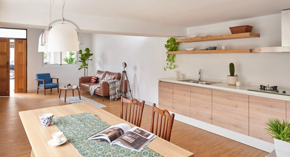 An open-concept kitchen and living space featuring light wood cabinetry, floating shelves, a dining table with pendant lights, and a leather sofa.