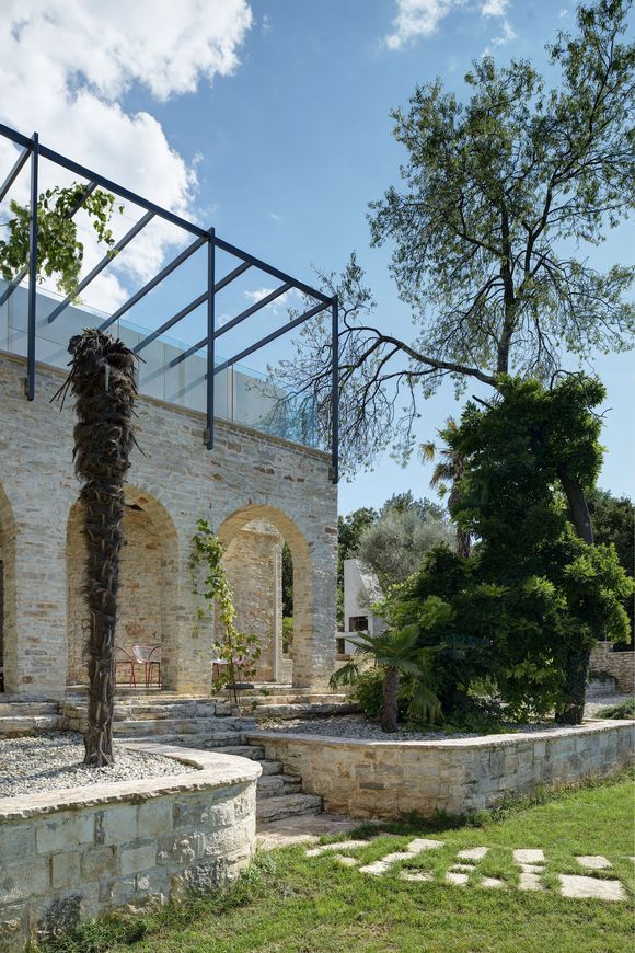 An outdoor stone patio features tall arched openings, a black metal pergola frame, and curved retaining walls surrounding garden beds.