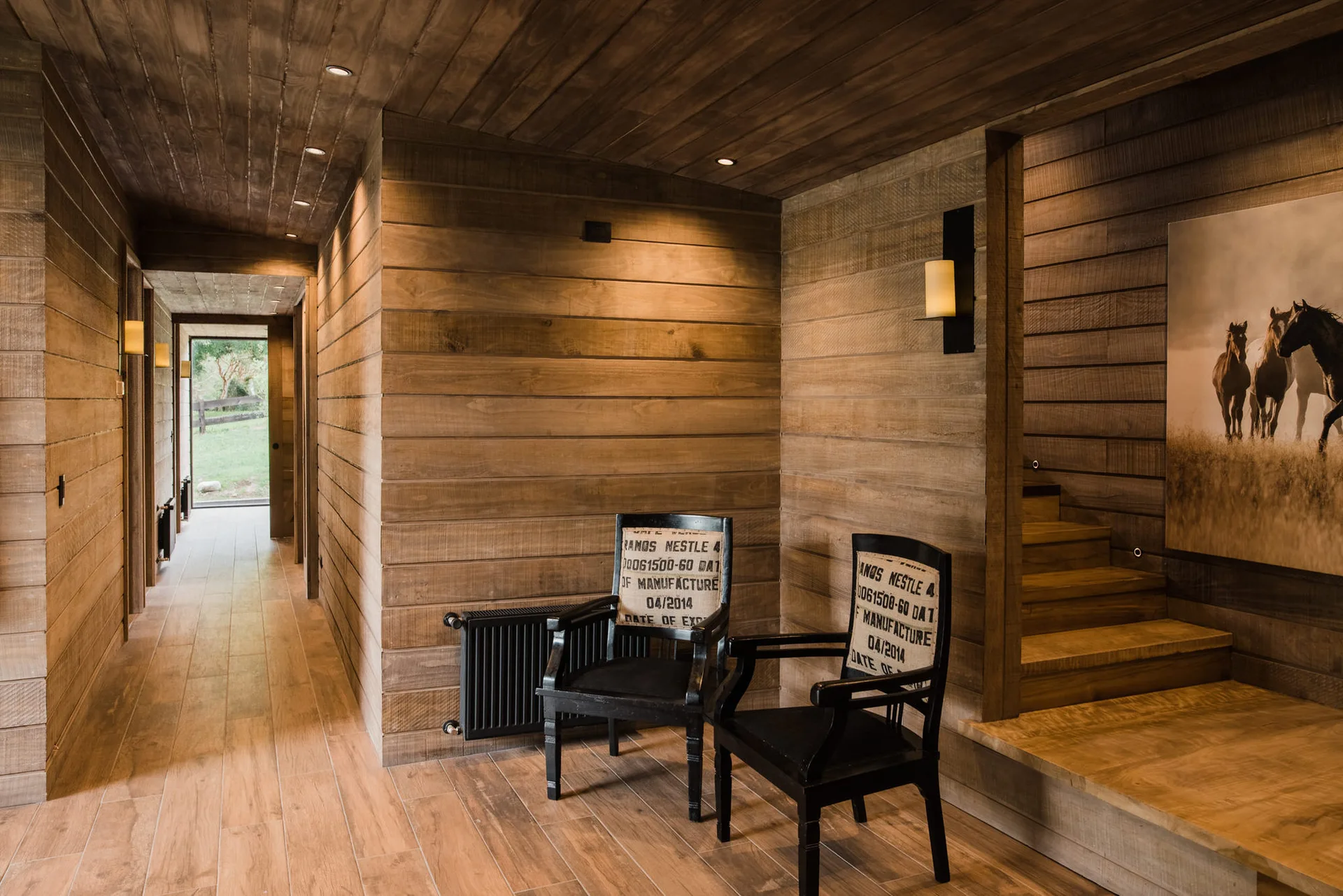 A wood-paneled hallway features horizontal timber cladding on the walls and ceiling, two black chairs, and a small wooden staircase.