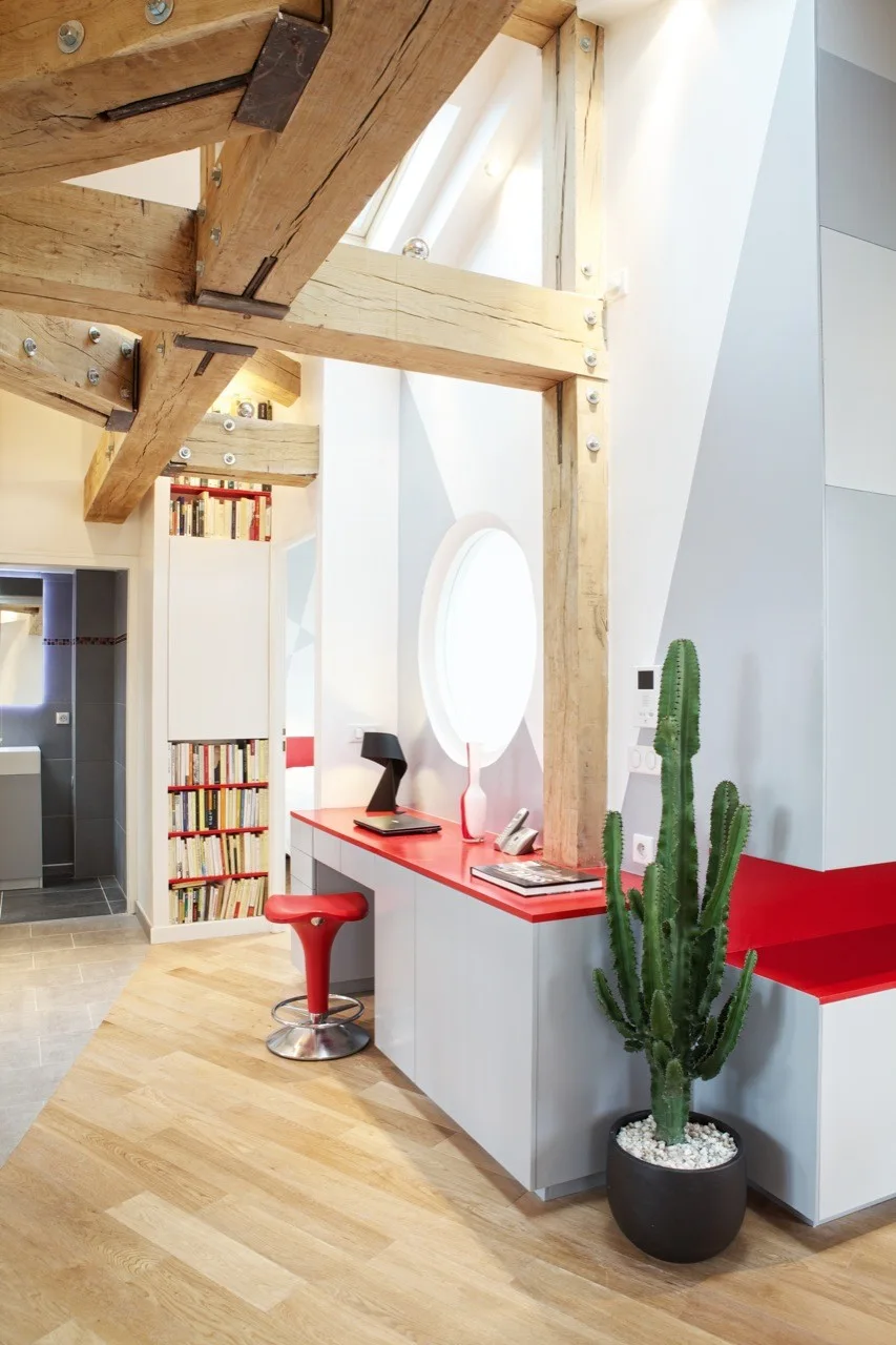 A light-filled workspace featuring rustic exposed timber beams, a built-in red desk with a matching red stool, and a circular window.
