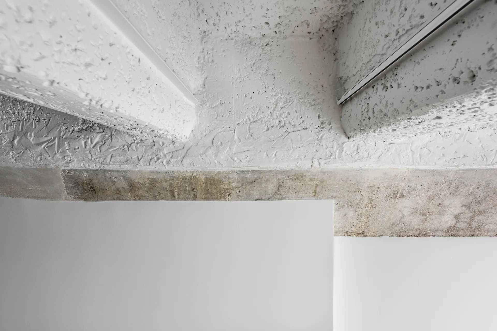 A rough white textured plaster ceiling meets an exposed grey concrete beam above a smooth painted wall with track lighting.