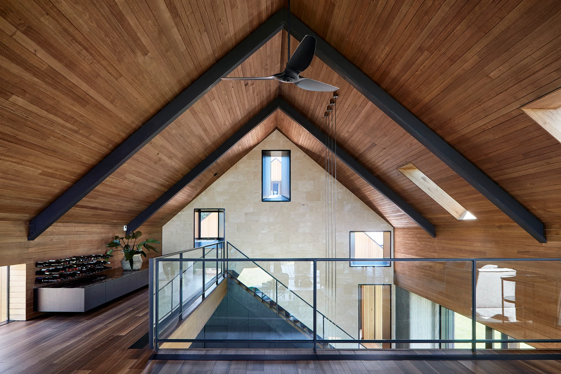 A modern residential loft landing features a vaulted wood-paneled ceiling, dark structural steel beams, a glass railing, and a stone tile back wall.