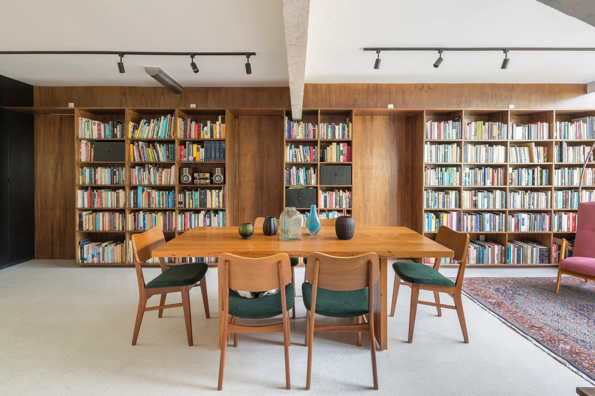 A wooden dining table and green-cushioned chairs stand before a massive wall of built-in wooden bookshelves in a room with a light terrazzo floor.