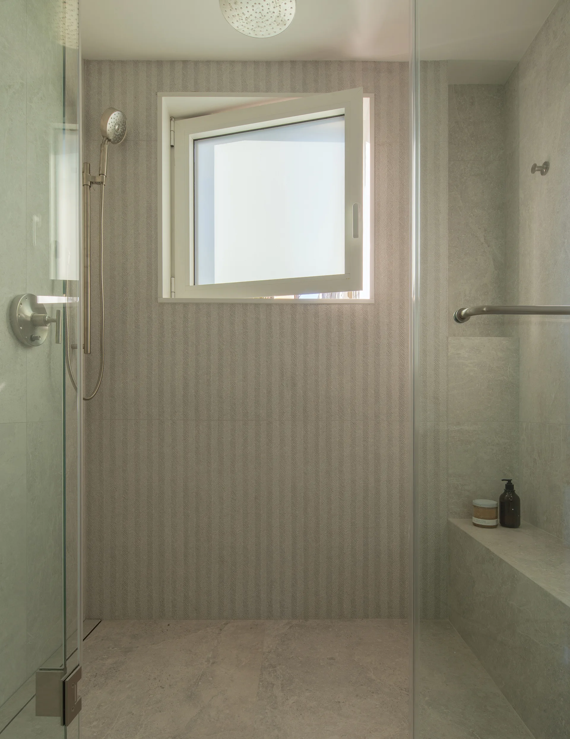 A modern glass shower enclosure with grey herringbone wall tiles, a built-in stone bench, and an open square window.