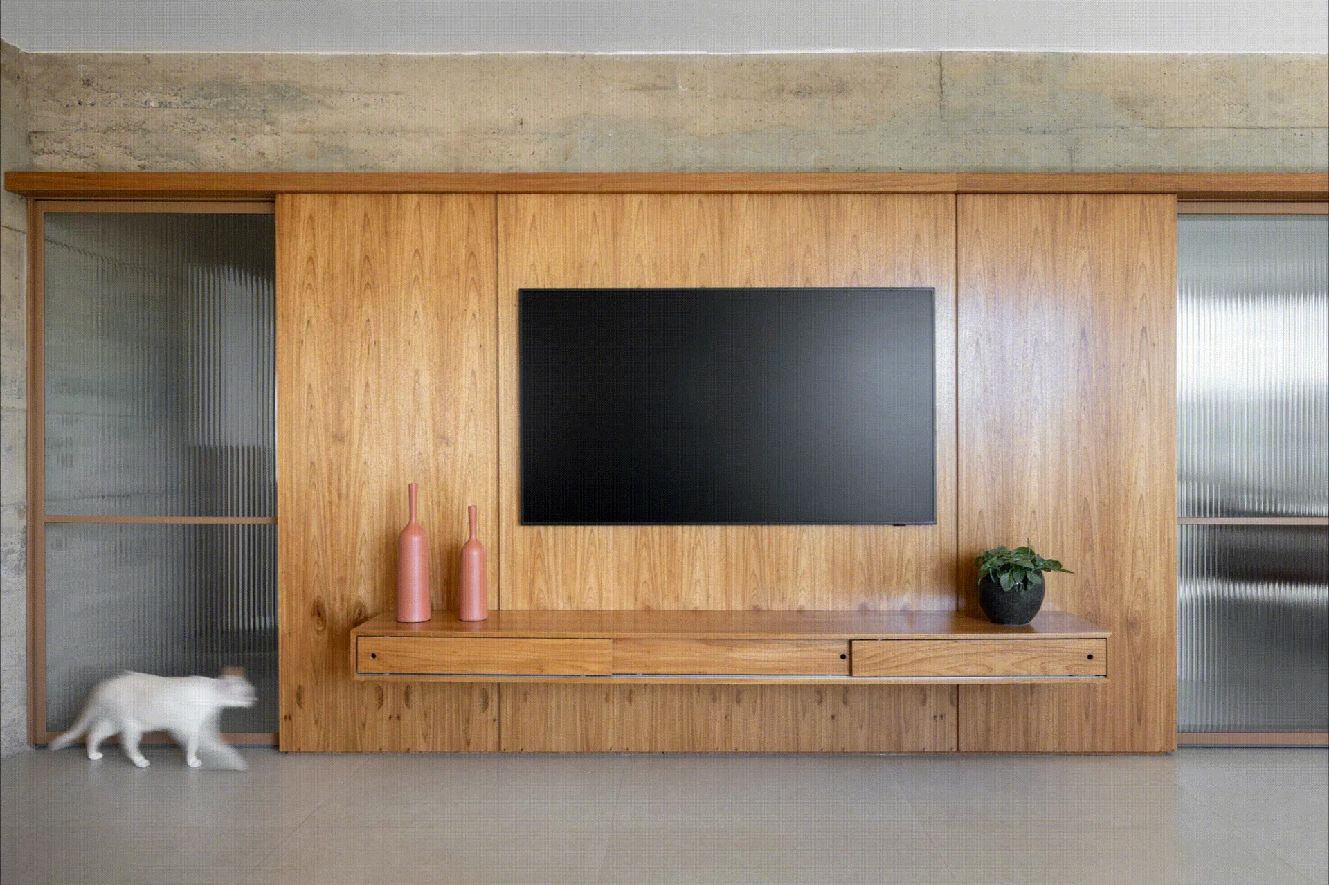 A flat-screen television is mounted on a warm wood-paneled wall above a floating media console, flanked by fluted glass sliding doors.