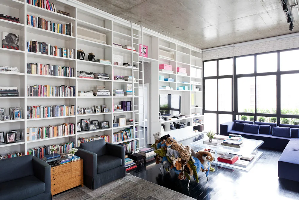 A spacious living room features a massive white built-in bookshelf with a library ladder, a navy sectional sofa, and a raw concrete ceiling.