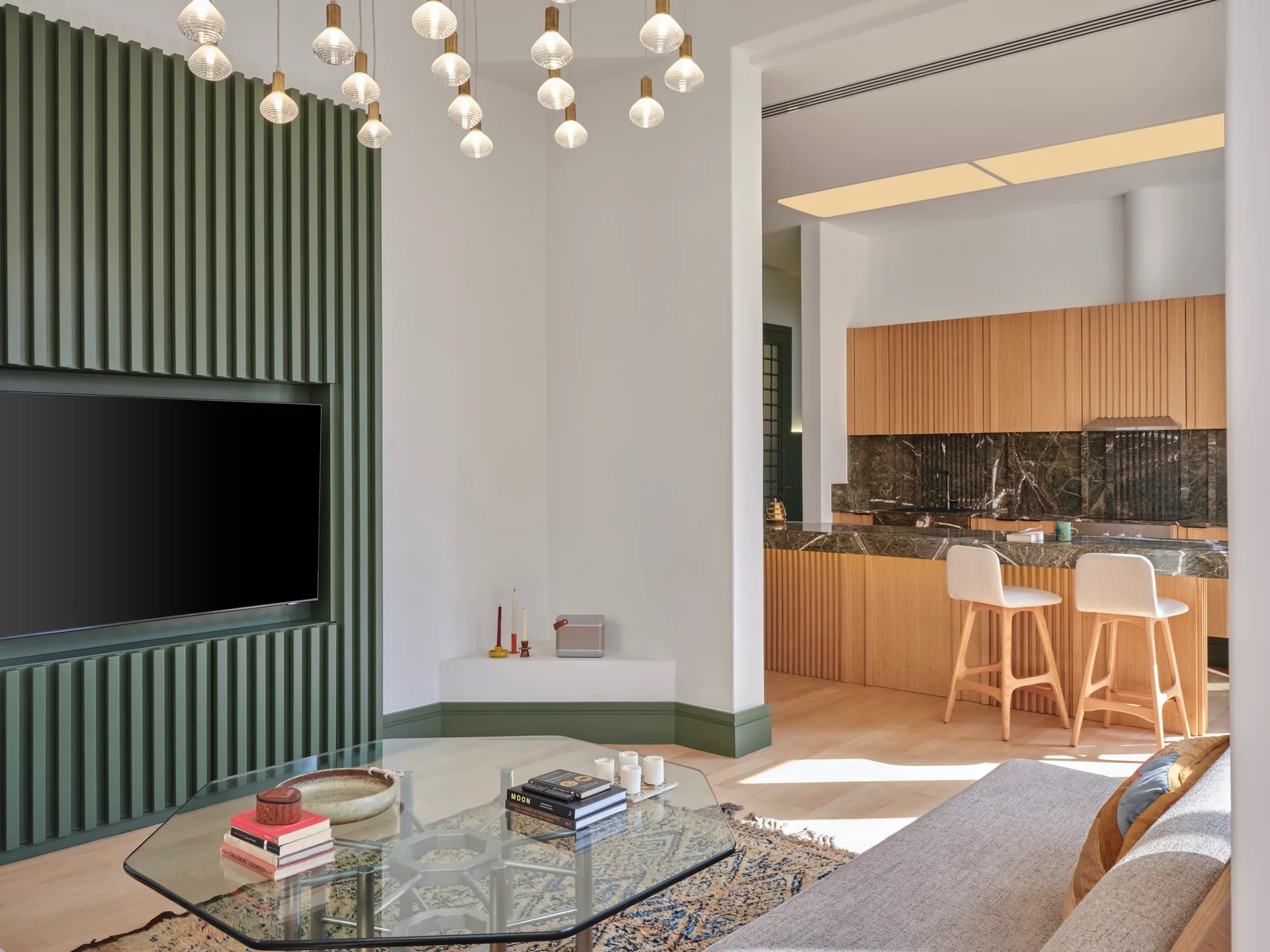 An open-concept living area features a green slatted feature wall with a TV, a glass coffee table, and a kitchen with light wood cabinets.