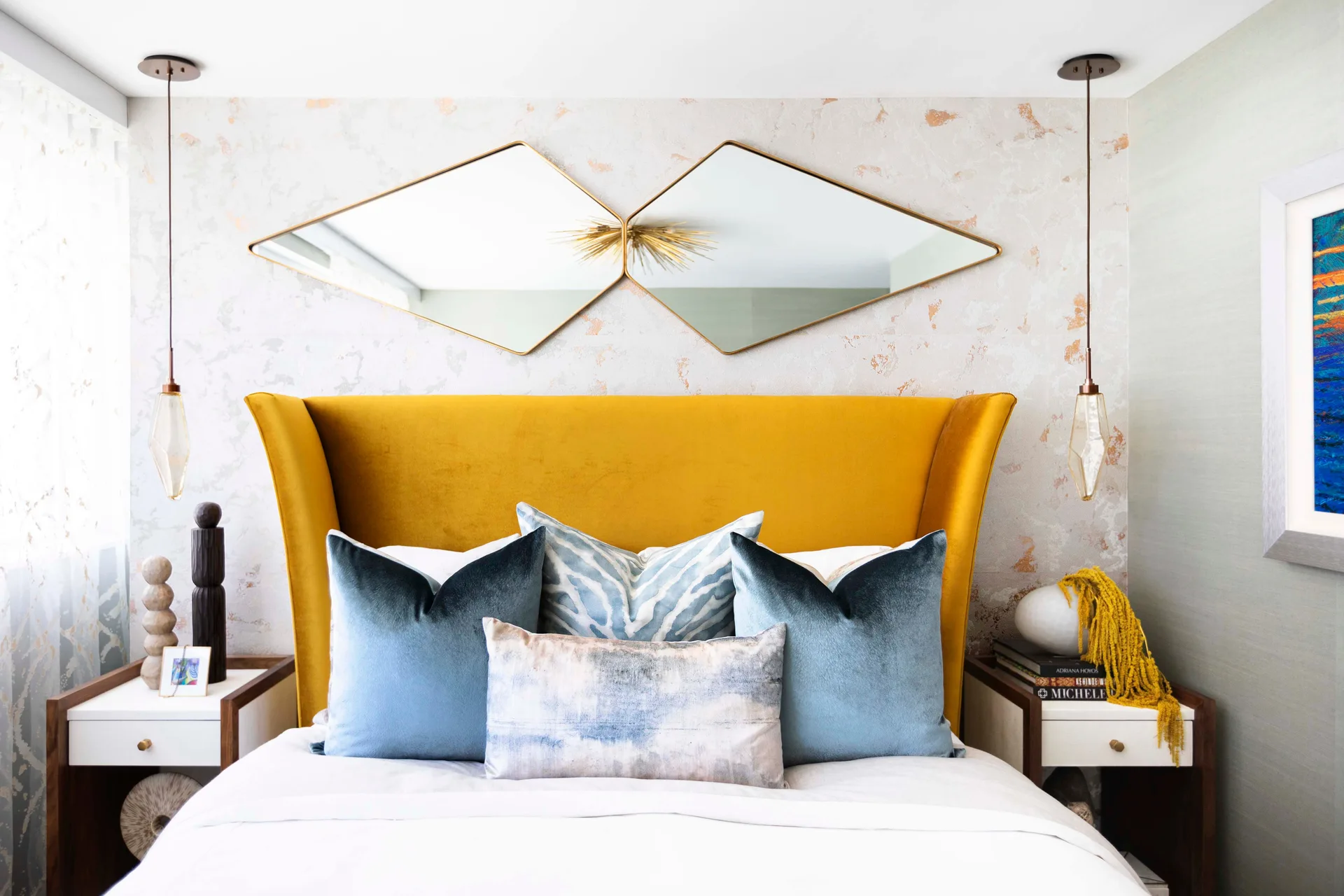 A bedroom features a bright yellow wingback headboard, blue throw pillows, and two diamond-shaped mirrors mounted side-by-side on patterned wallpaper.