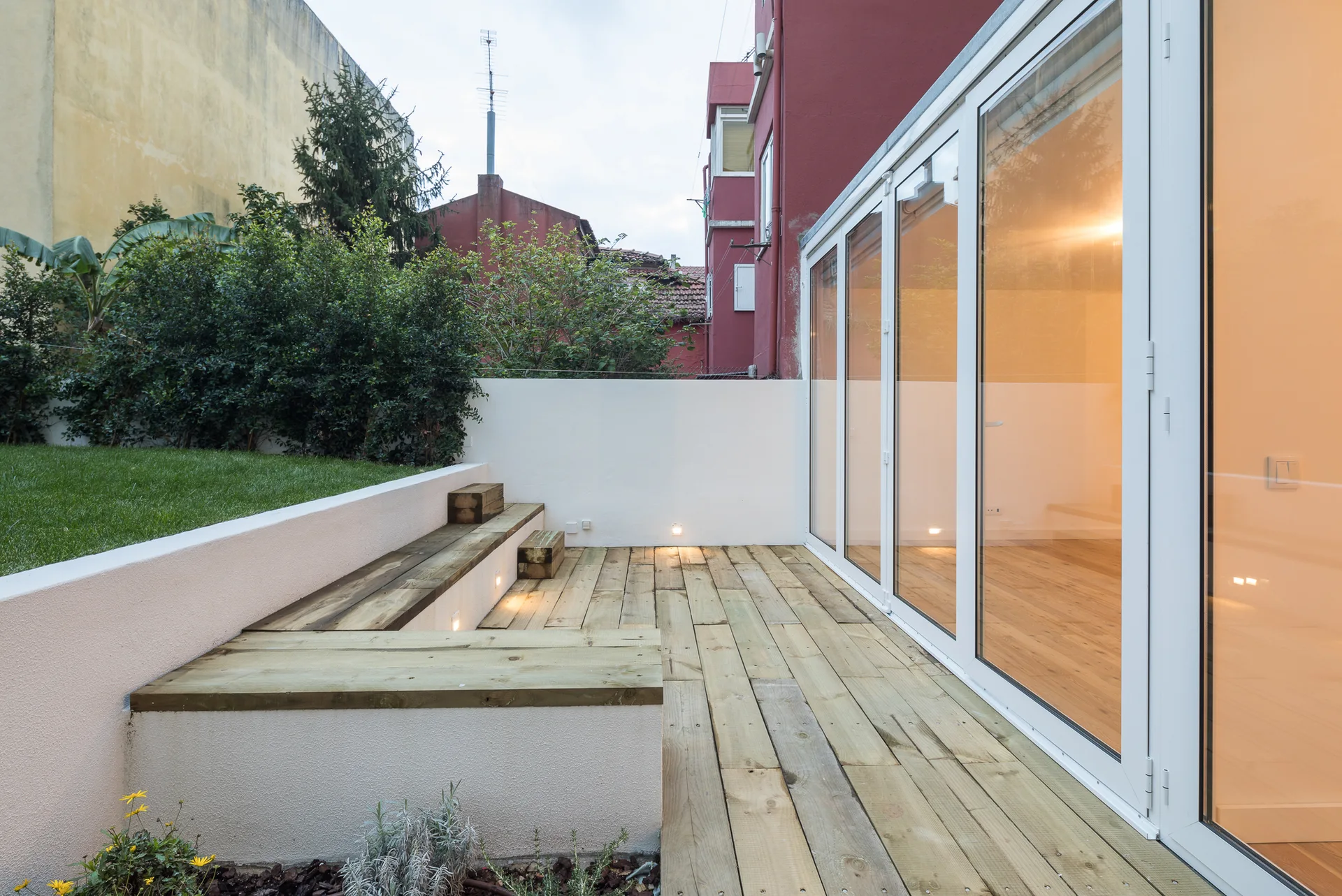 A wooden patio deck with integrated steps, bench seating, low recessed lighting, and large glass doors leading inside.