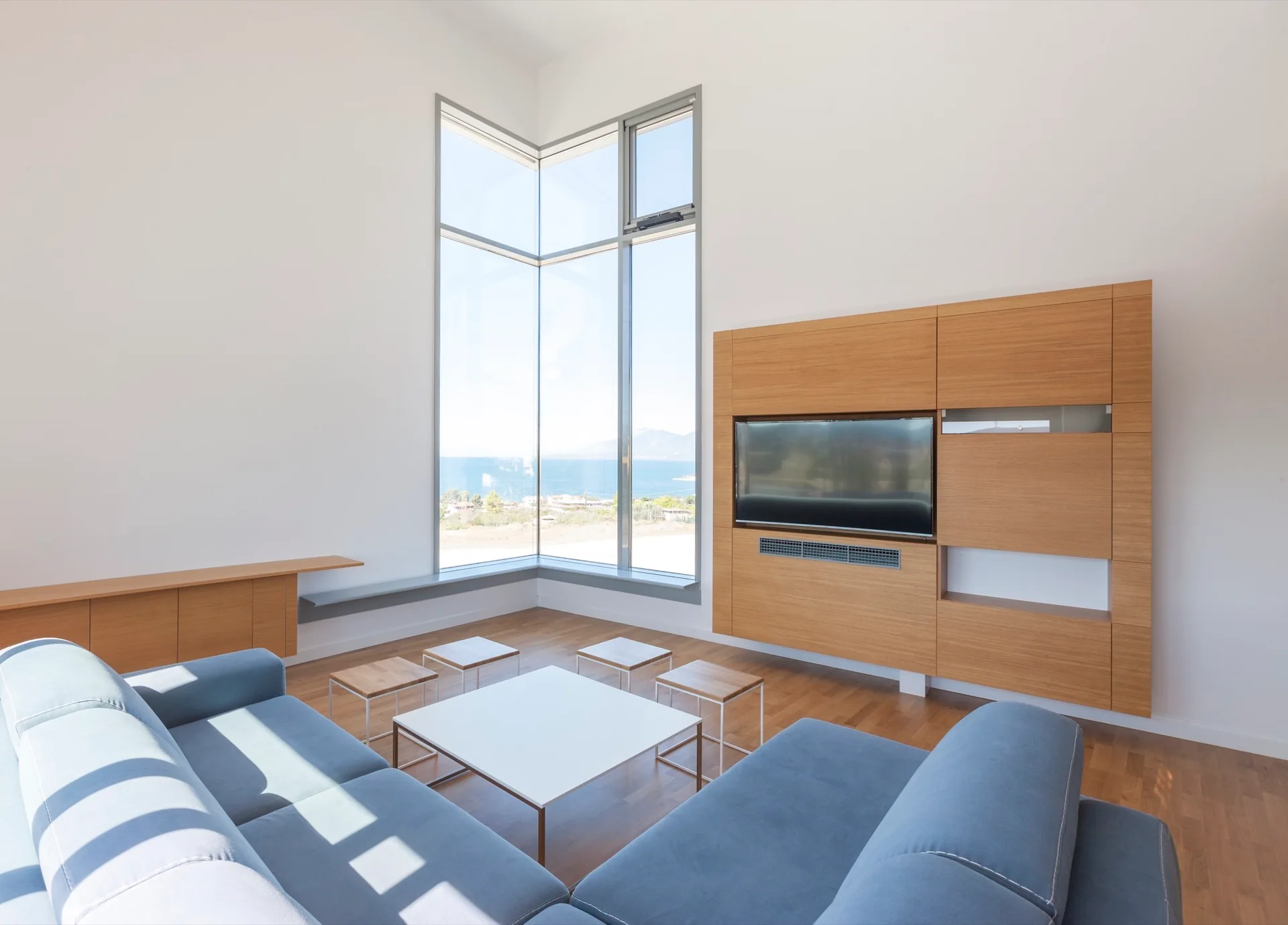 A minimalist living room features a blue modular sofa, a white coffee table, a wood media console, and a tall corner window with a coastal view.