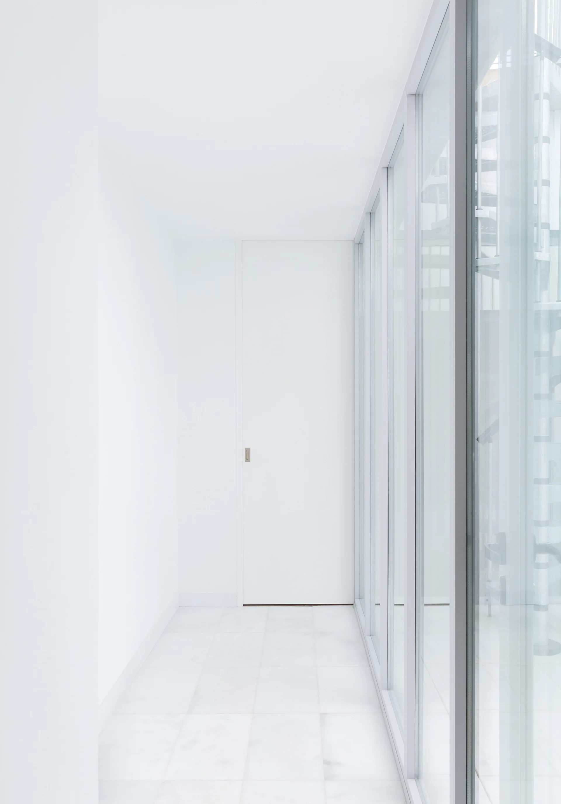 A bright white minimalist hallway featuring a flush-fit door, stone tile flooring, and a glass partition looking onto a spiral staircase.