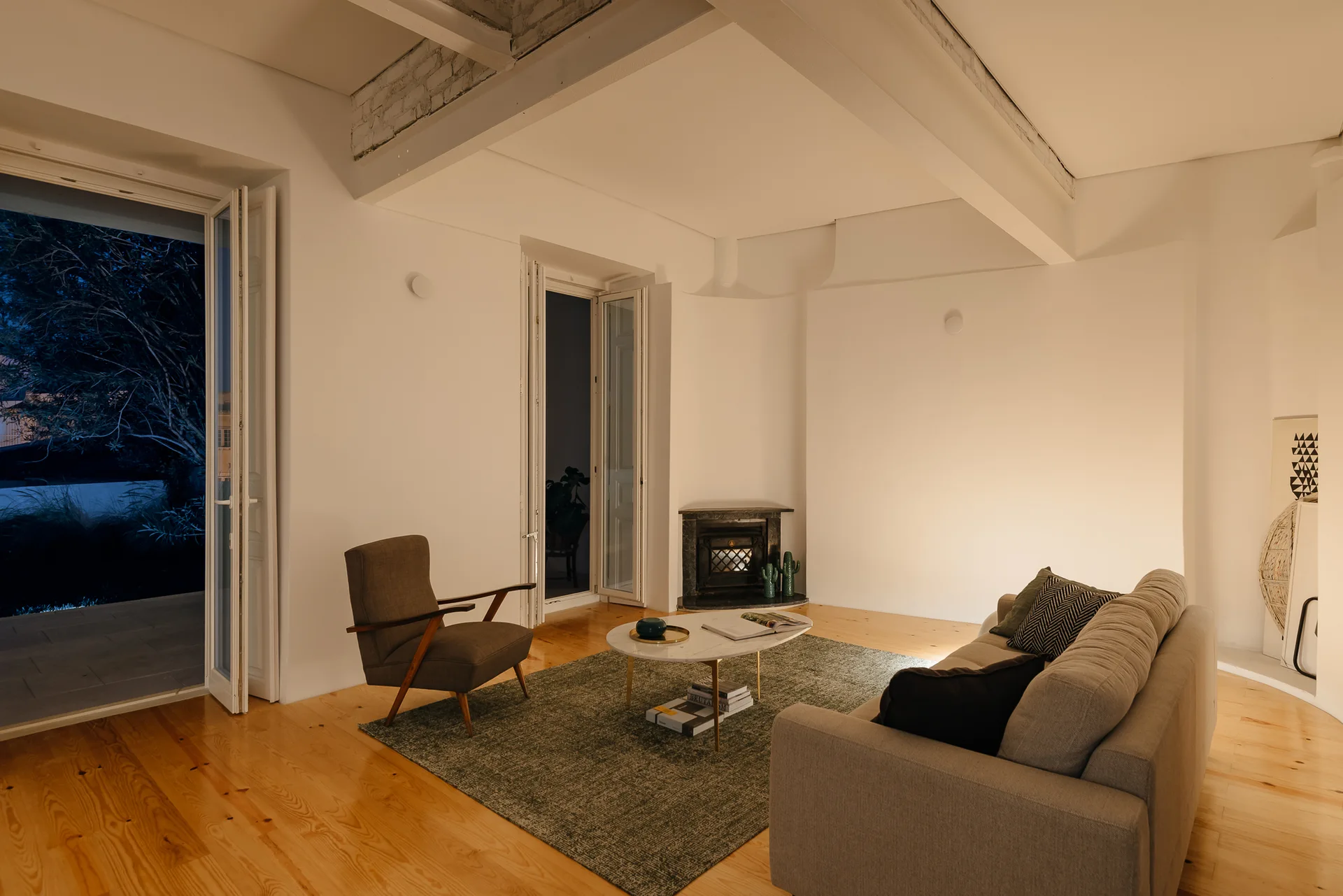 A warm-lit living room features a grey sofa, a mid-century armchair, and an oval marble coffee table on a green rug beneath white ceiling beams.