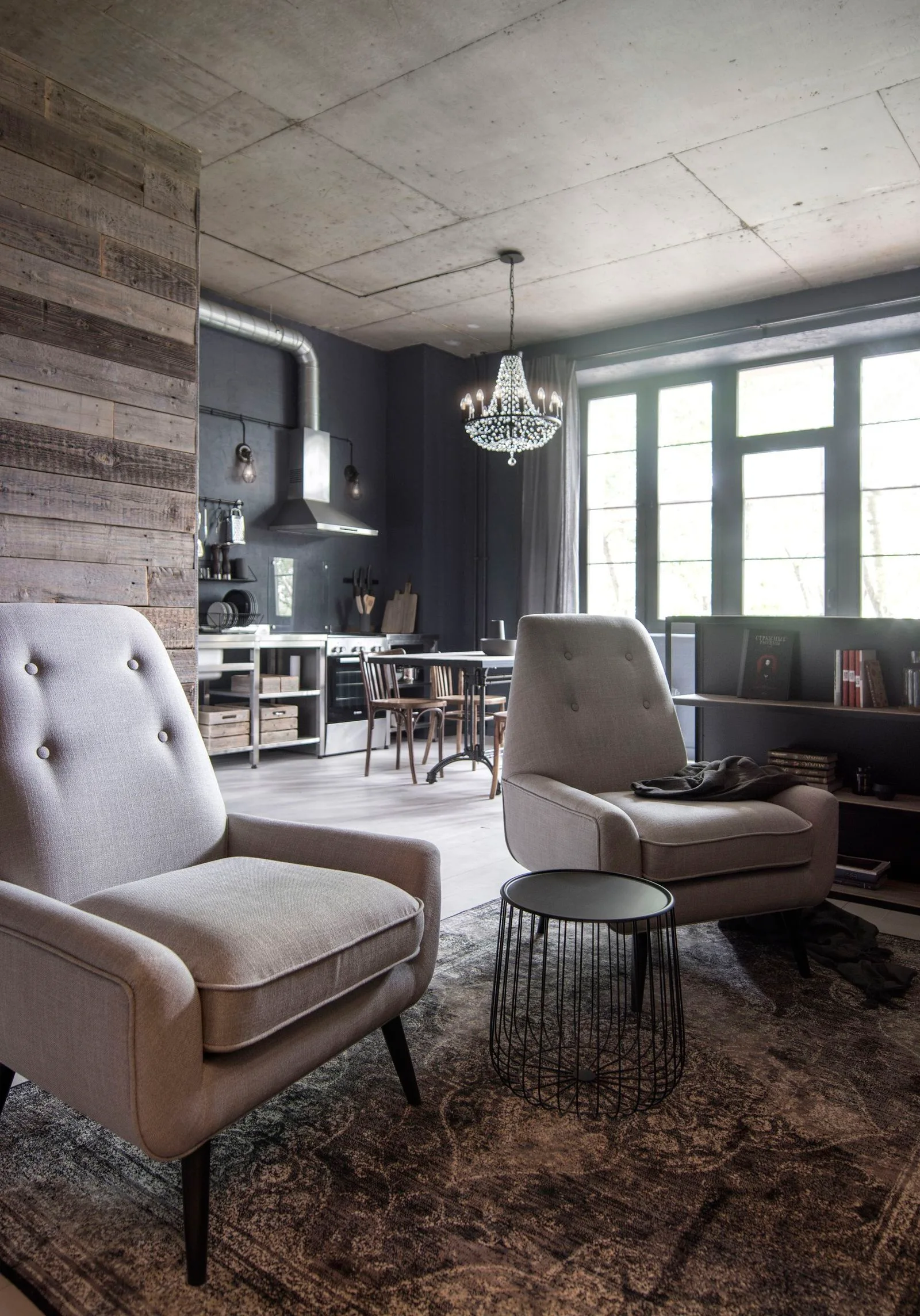 Two grey tufted armchairs sit on a distressed rug in an industrial living room featuring a raw concrete ceiling and a background metal kitchen.