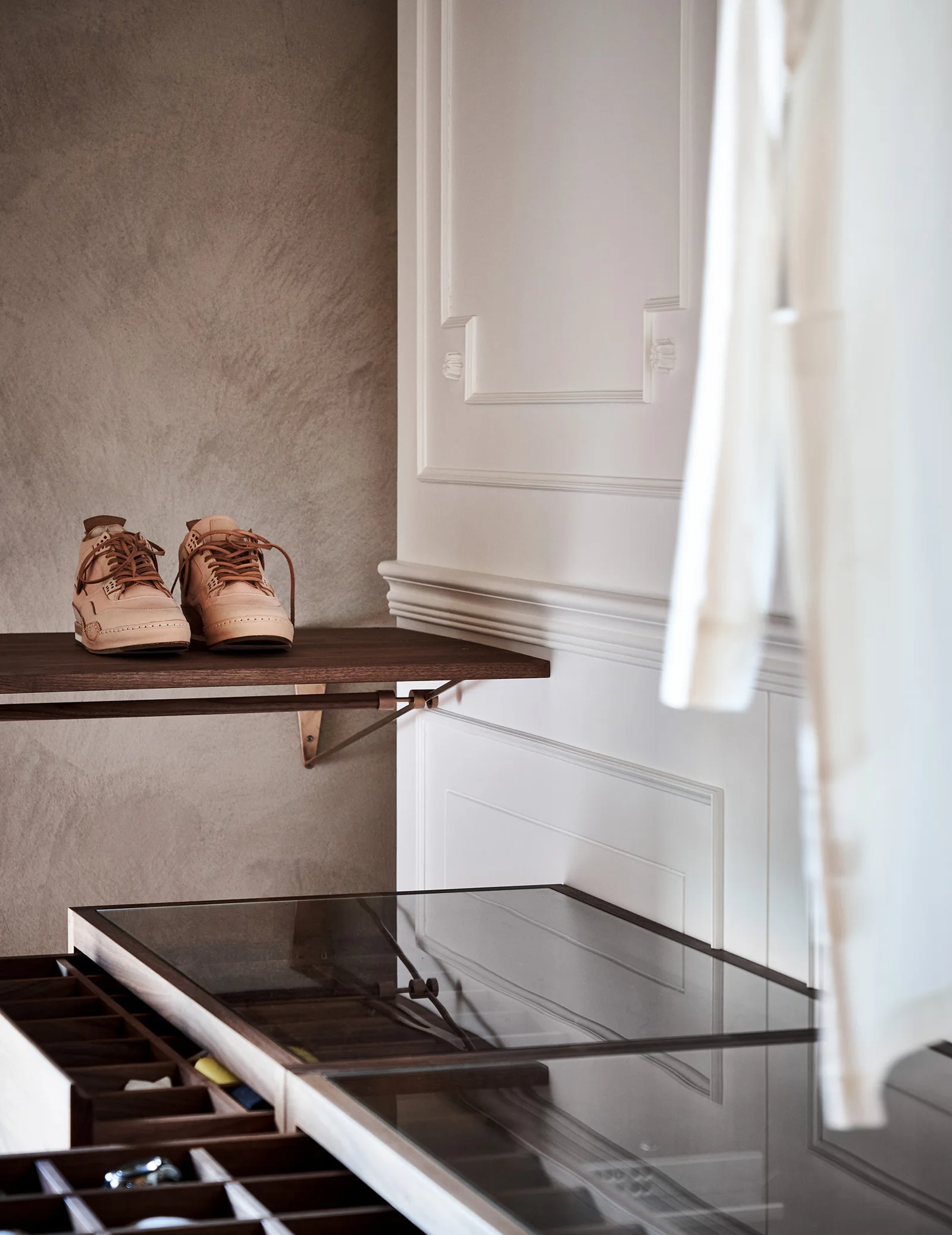 A custom closet features dark wood drawers with a glass top, a wooden display shelf with leather sneakers, and white paneled walls.