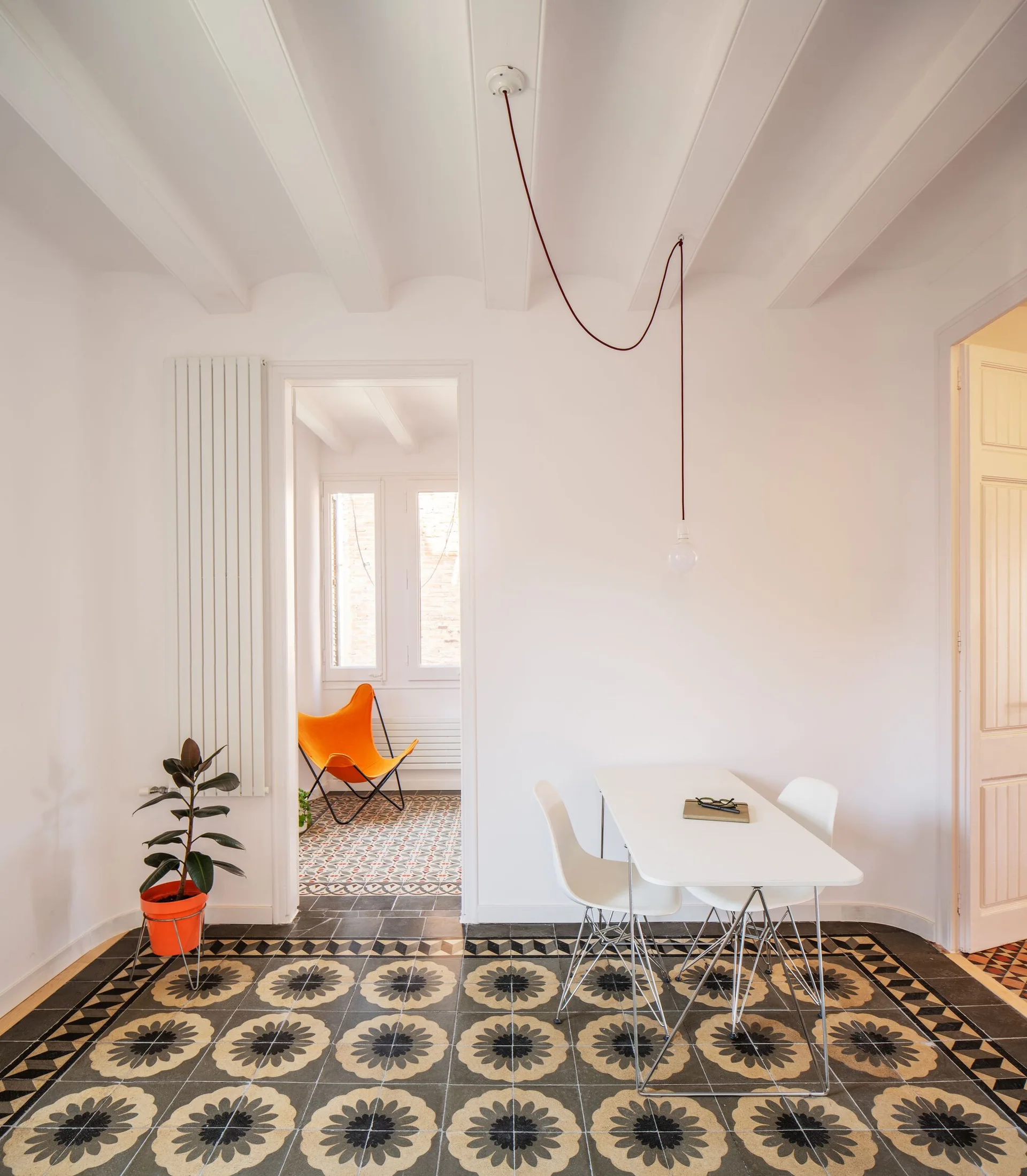 A minimalist white dining table and chairs set on a bold black and grey patterned tile floor beneath white exposed ceiling beams.