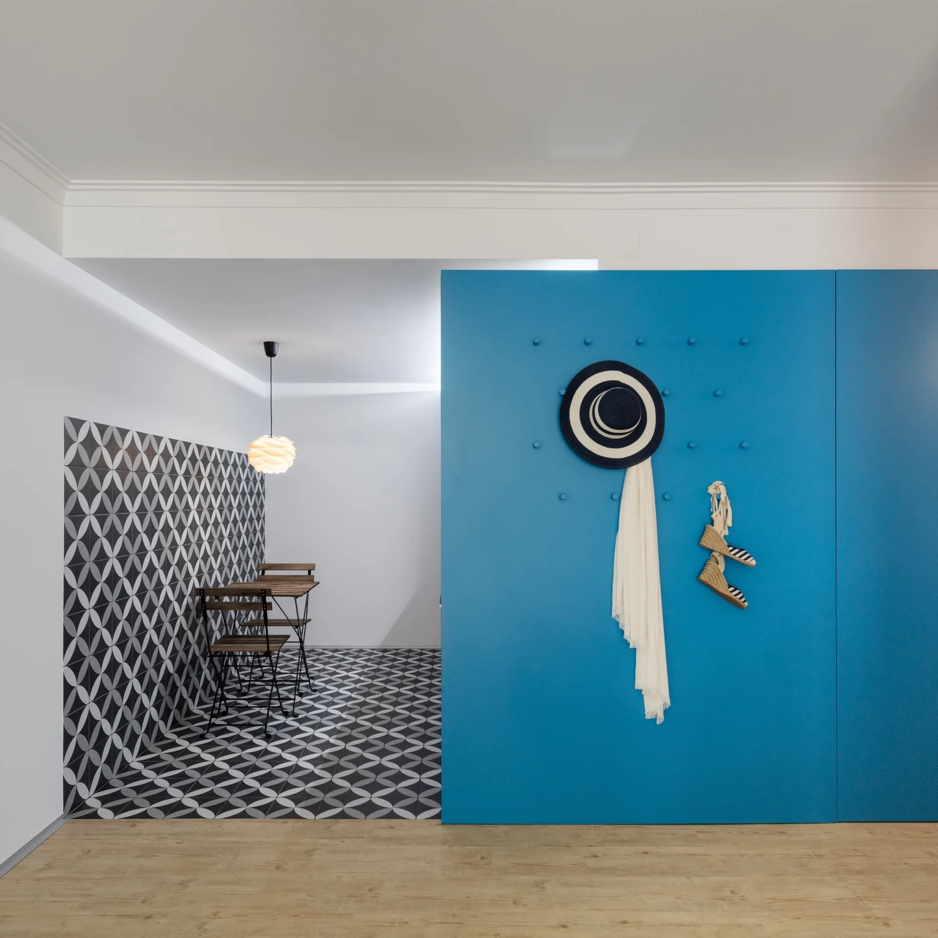 A bright blue partition wall with hanging pegs stands next to a dining area featuring geometric black and white patterned tiles and a wooden bistro set.