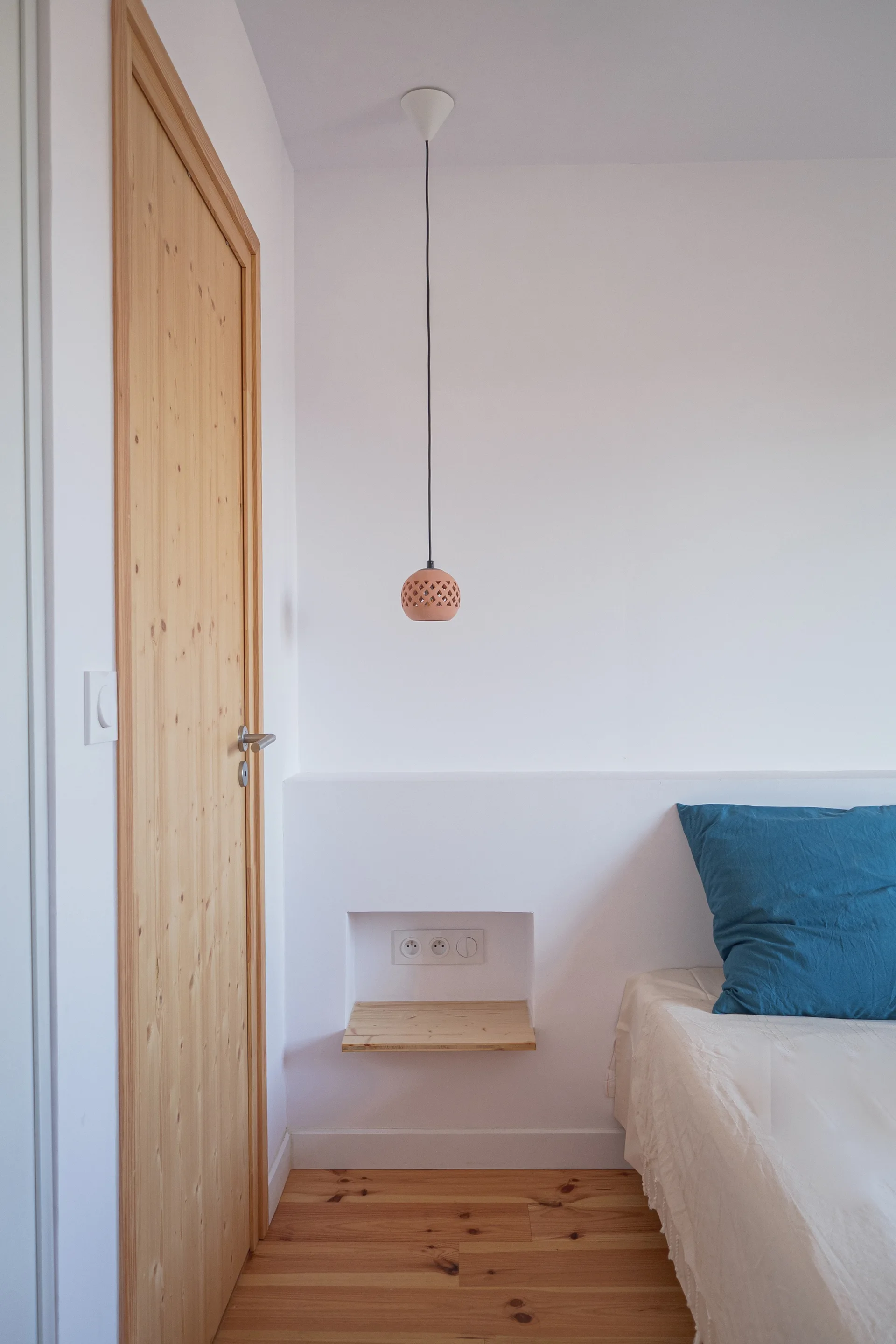 A minimalist bedroom corner features a natural wood door, a recessed wall niche with a small wood shelf, and a hanging pendant light next to a bed.