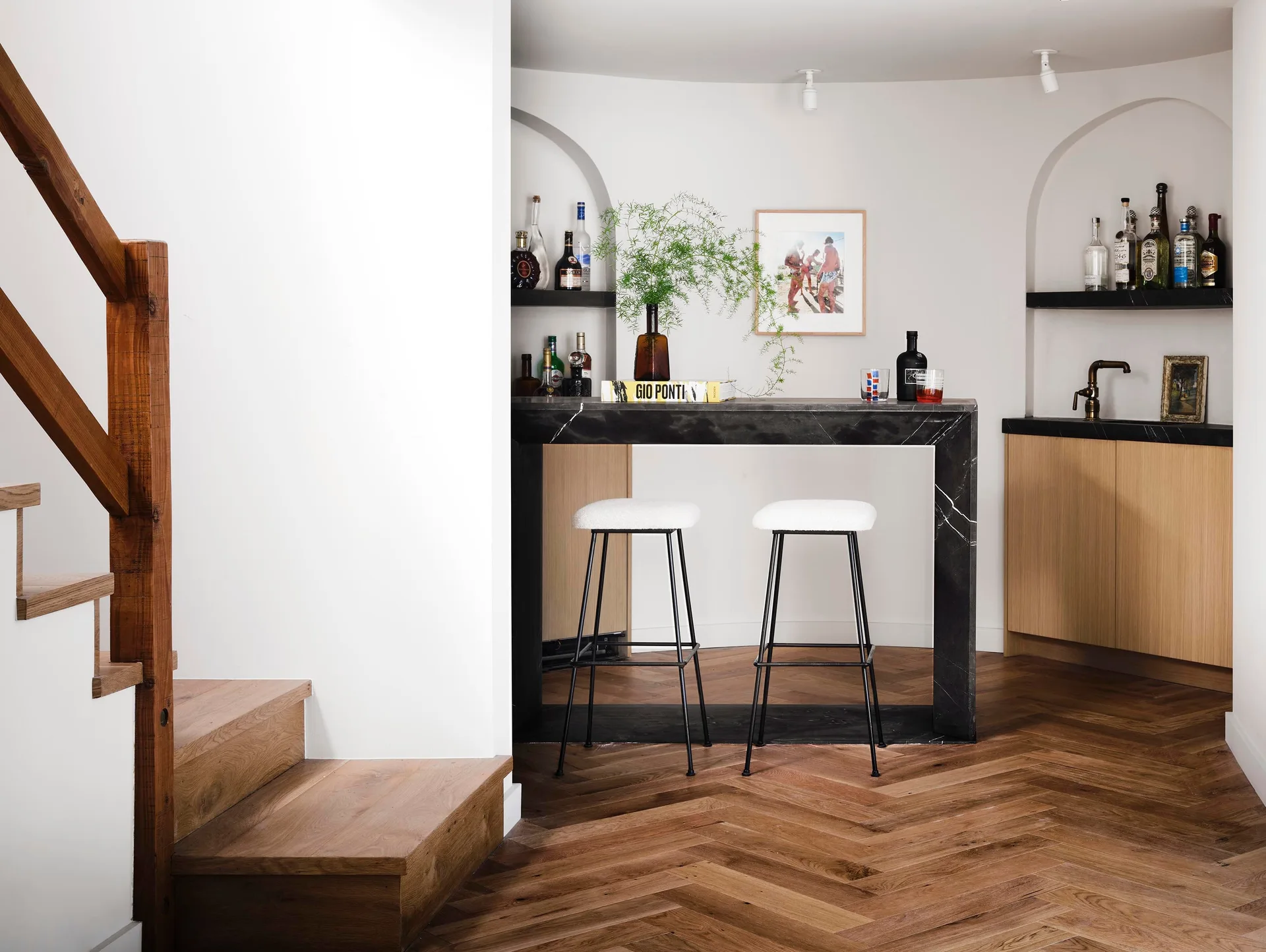 A modern home bar featuring a black stone counter, two white-cushioned bar stools, arched wall niches, and herringbone wood flooring.