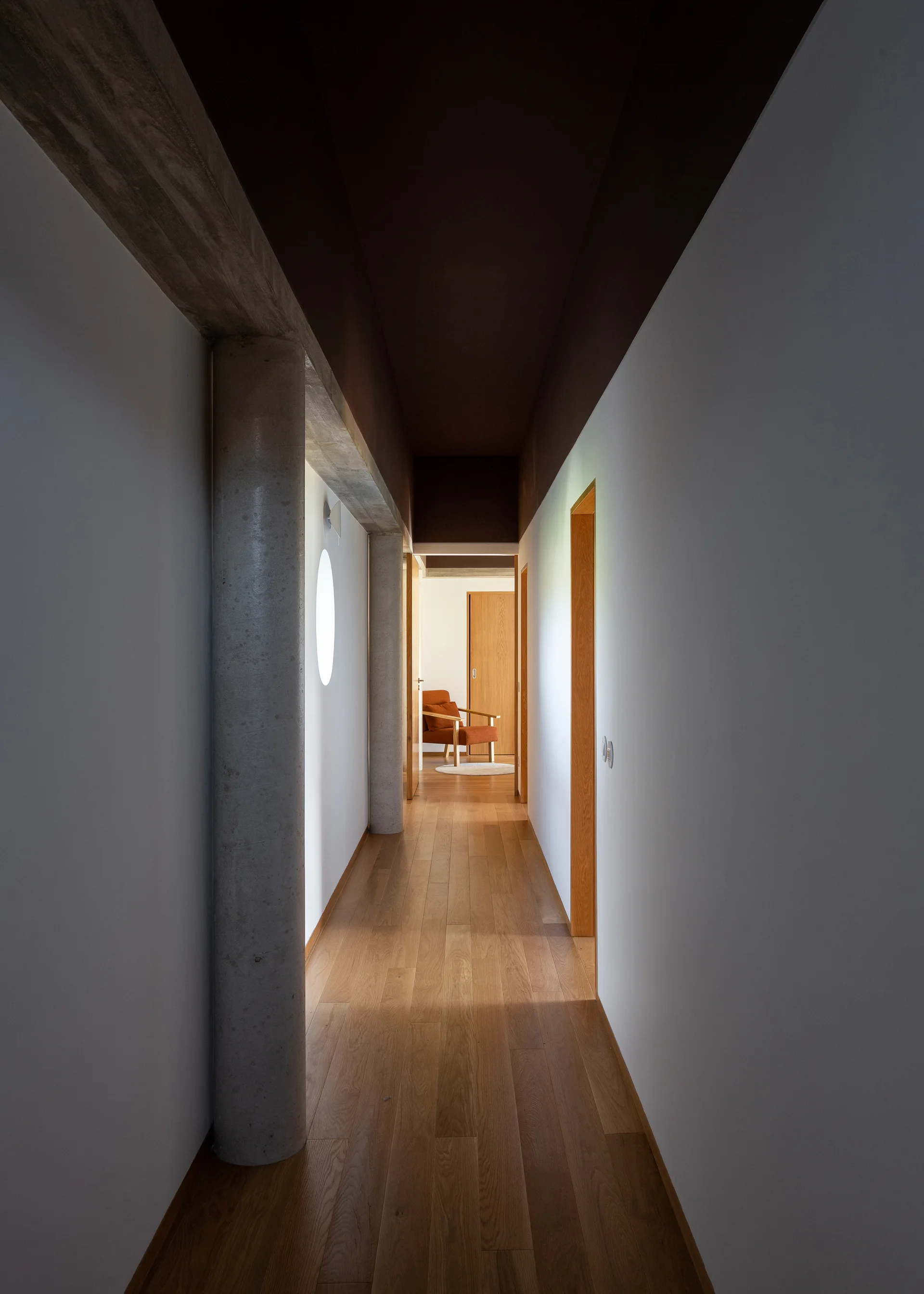 A minimalist hallway features cylindrical concrete columns, a circular window in a white wall, wood flooring, and a dark recessed ceiling.