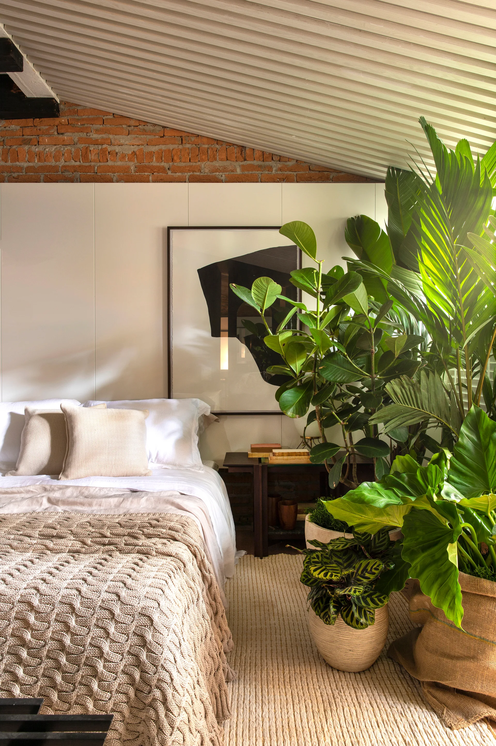 A bedroom featuring a bed with a beige knit blanket, a dense cluster of large green potted plants, an exposed brick wall, and a sloped ceiling.