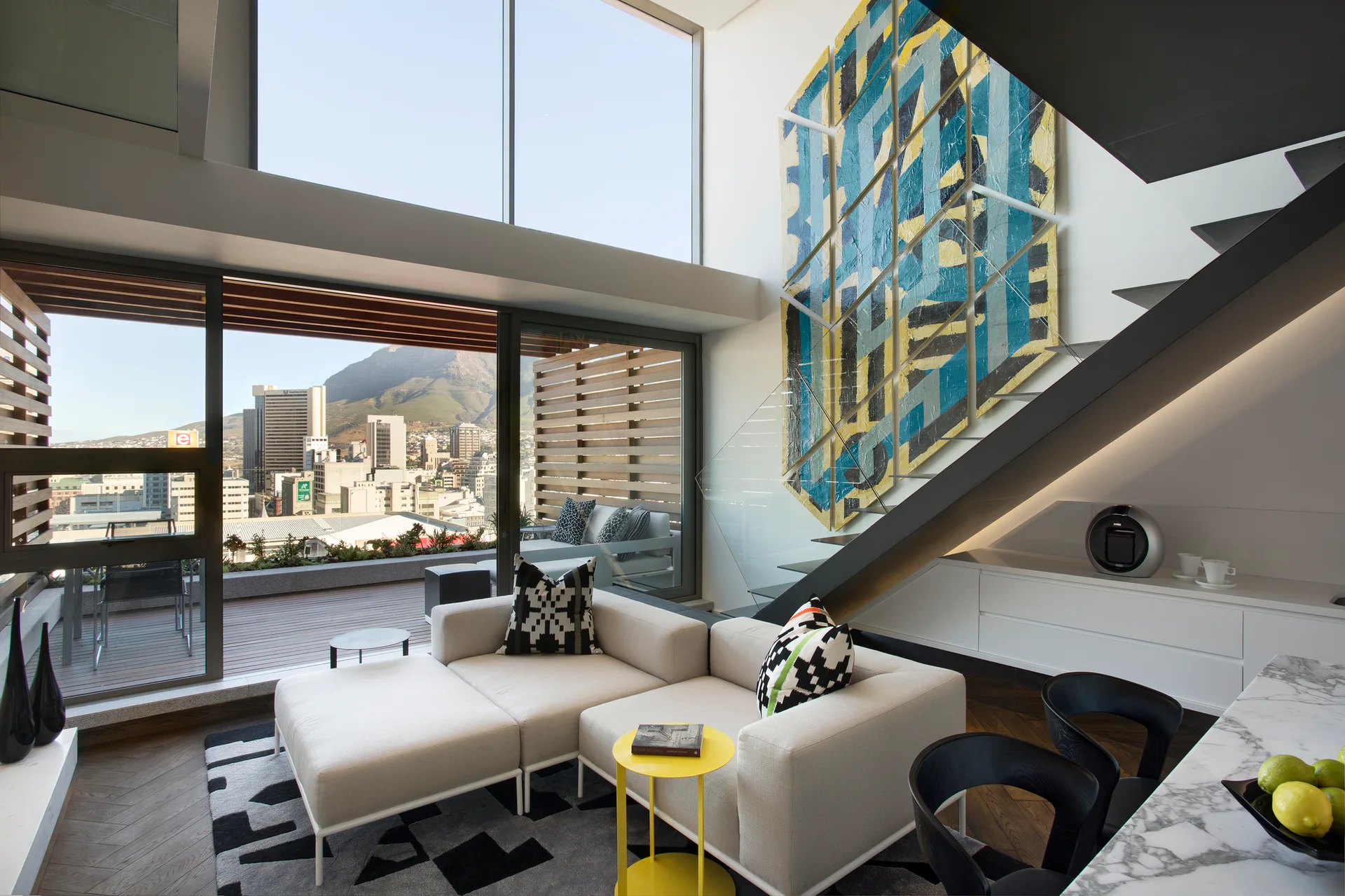 A modern double-height living room featuring a cream sectional sofa, a black-and-white geometric rug, and large abstract wall art next to a staircase.