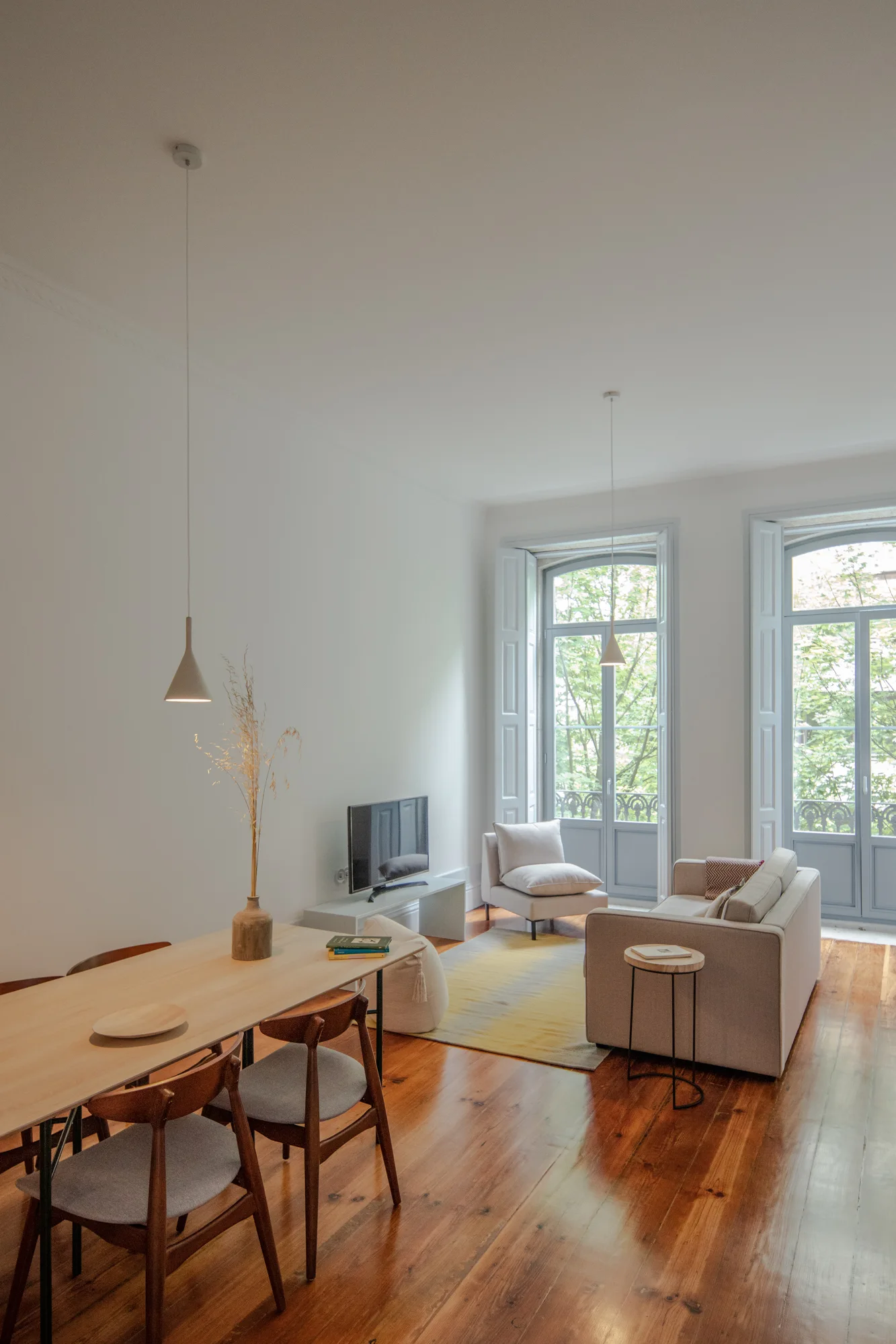 A light-filled open-concept living room features a wooden dining table, a beige sofa on a yellow gradient rug, and pale blue French doors.