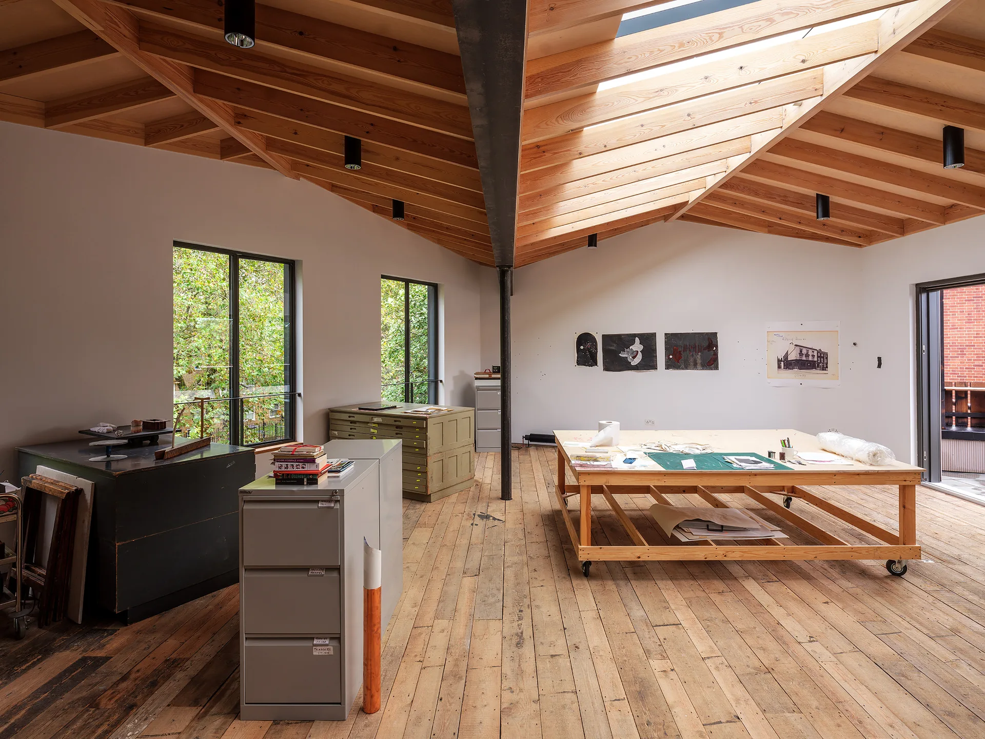 An open artist studio featuring a vaulted wood ceiling, exposed timber beams, a large mobile wooden work table, and metal filing cabinets.
