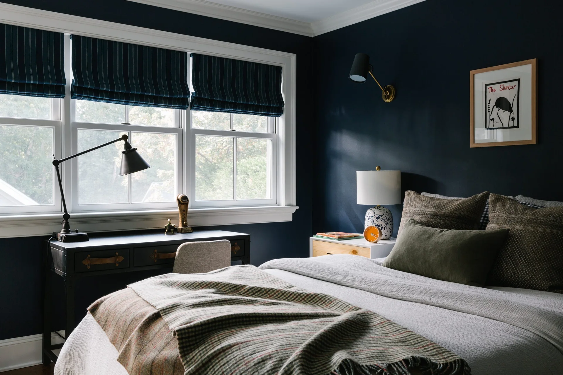 A bedroom with dark navy walls, a bed layered with textured blankets, and a black desk positioned beneath a window with striped roman shades.