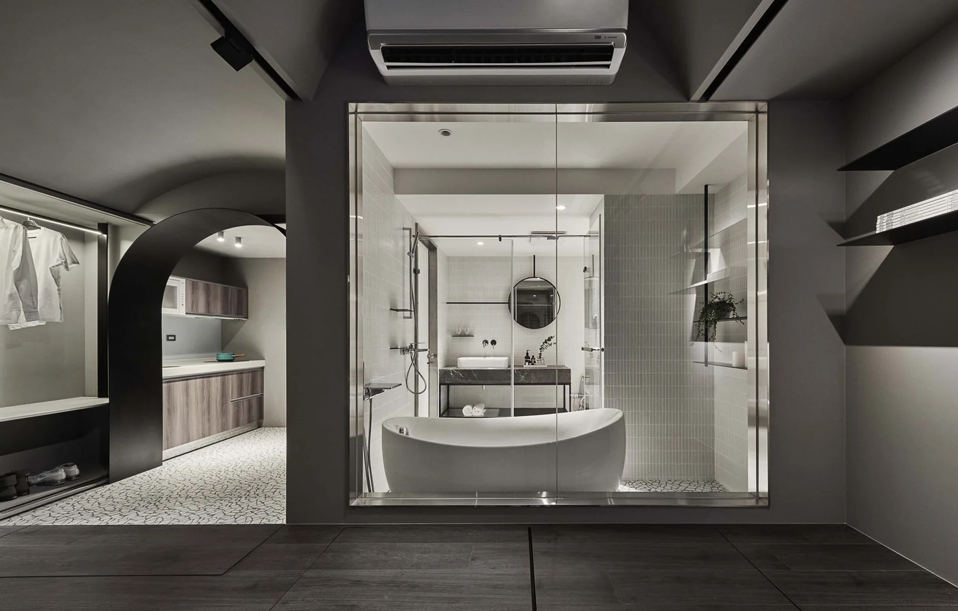 A modern glass-enclosed bathroom with a freestanding white bathtub sits next to a dark archway leading to a kitchen.