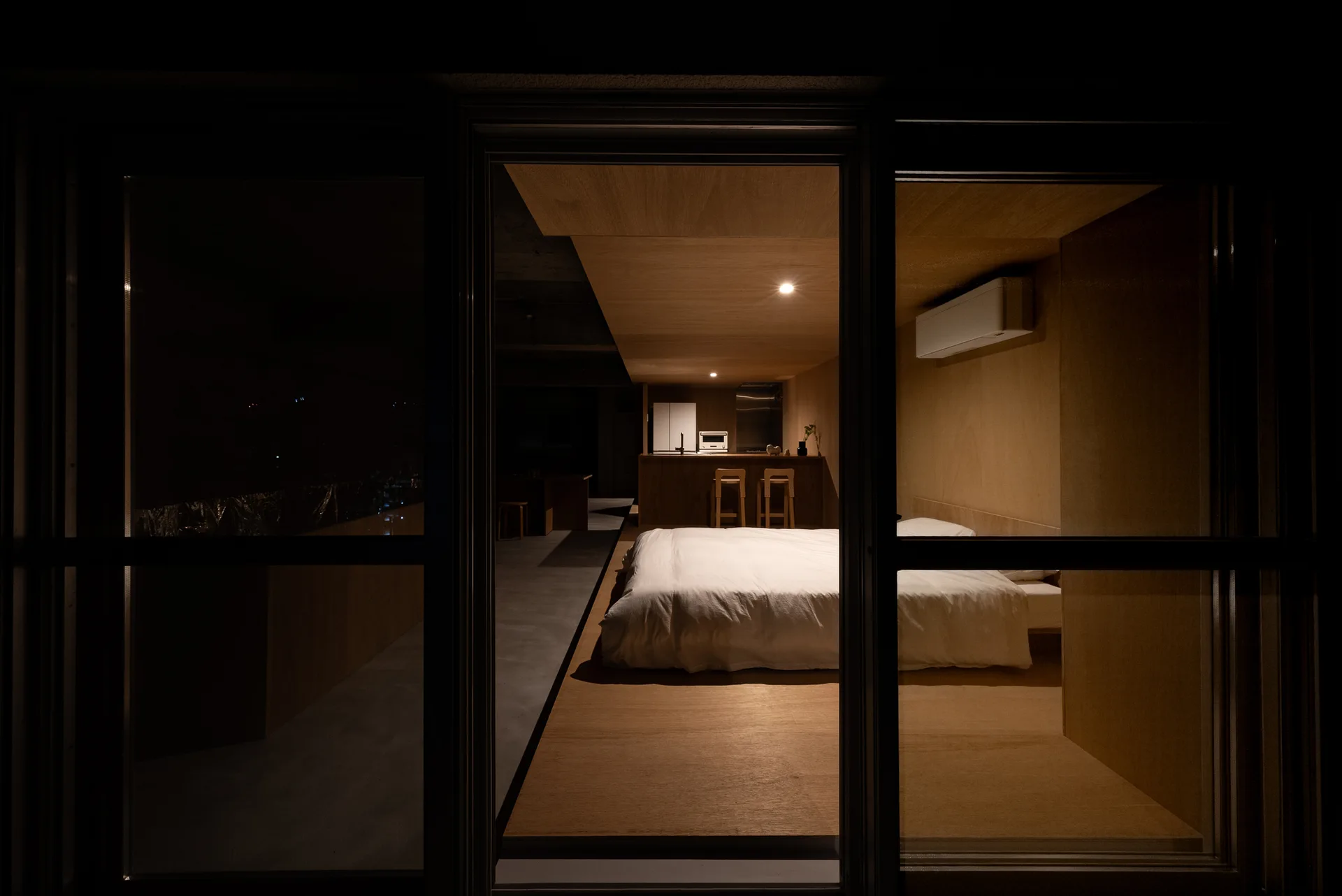 A view through a glass partition into a minimalist wood-paneled bedroom featuring a bed on a raised platform at night.