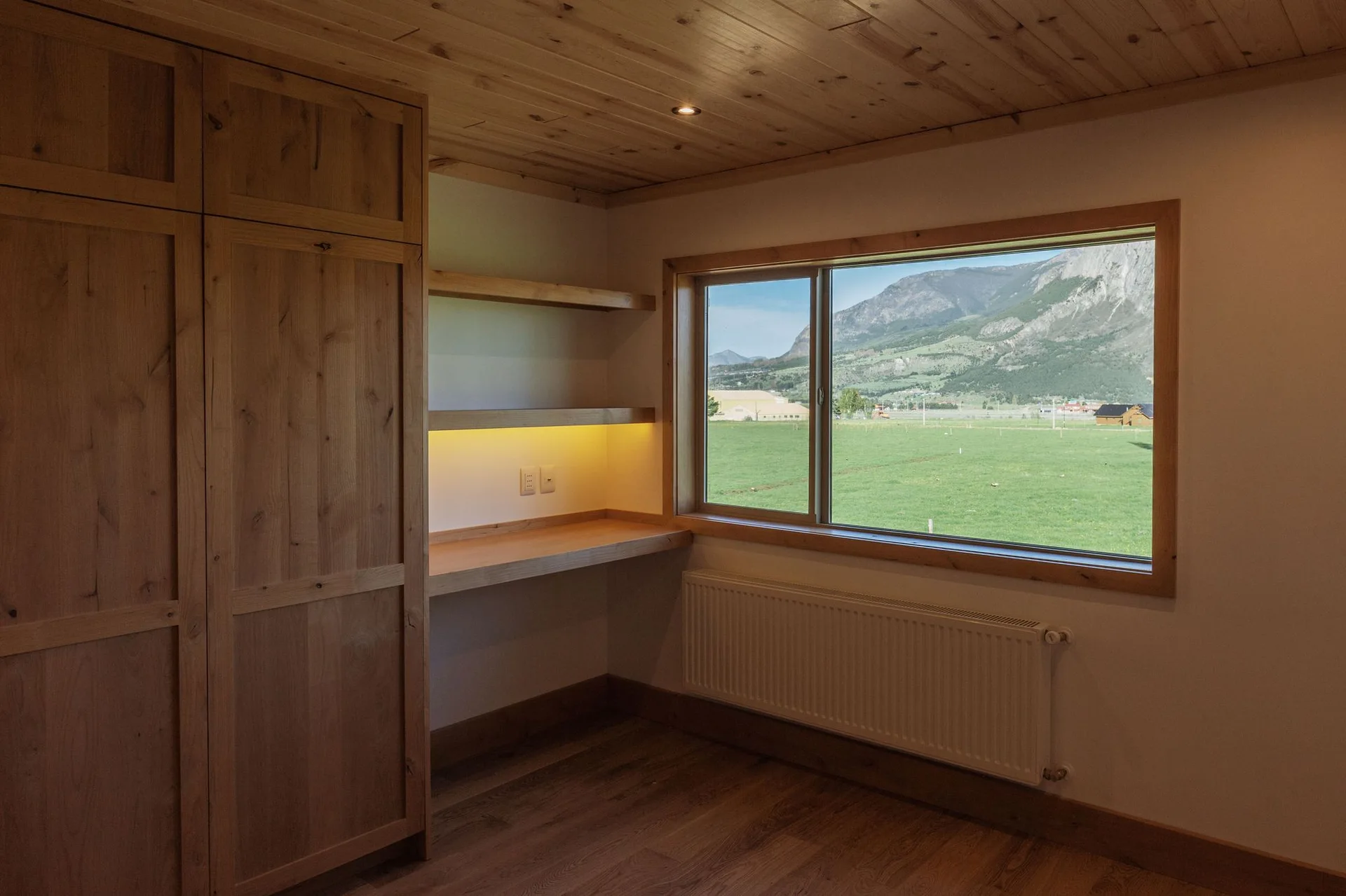 A minimalist home office features built-in wood cabinets, a floating desk with under-shelf lighting, and a large window looking out at green hills.
