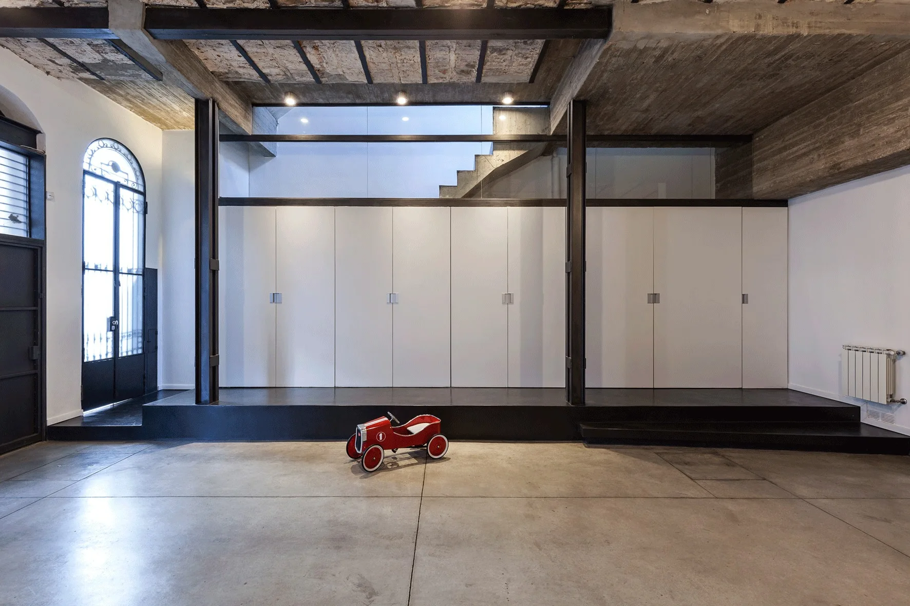 An industrial loft featuring a concrete floor and ceiling, a large wall of white built-in storage on a black platform, and a small red toy car.