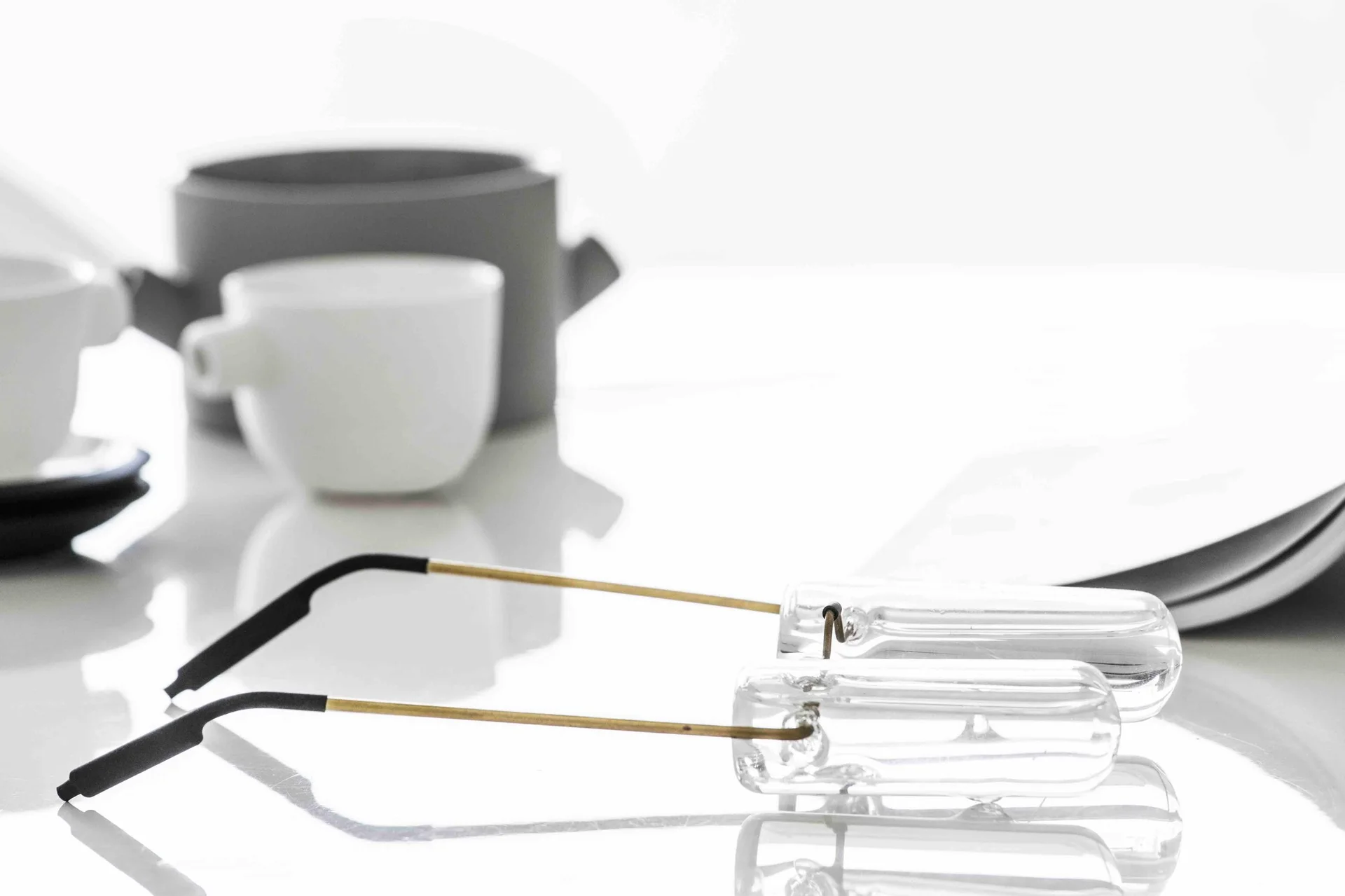 A pair of clear frame eyeglasses with thin metal arms rests on a glossy white table near ceramic cups and a gray teapot under bright light.