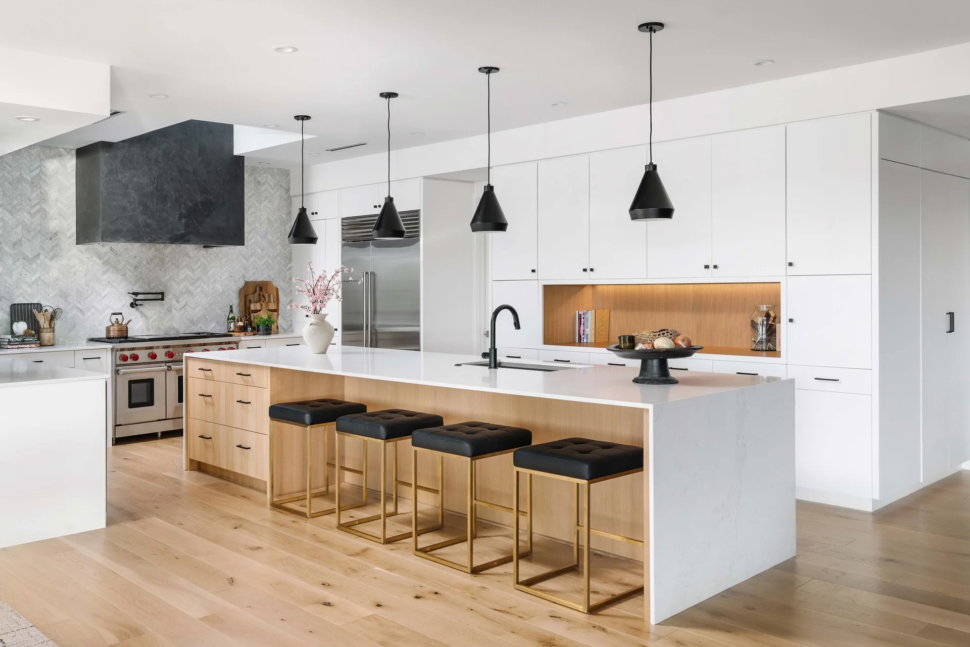 A modern kitchen features a large wood-faced island with black bar stools, a herringbone tile backsplash, and black pendant lights.