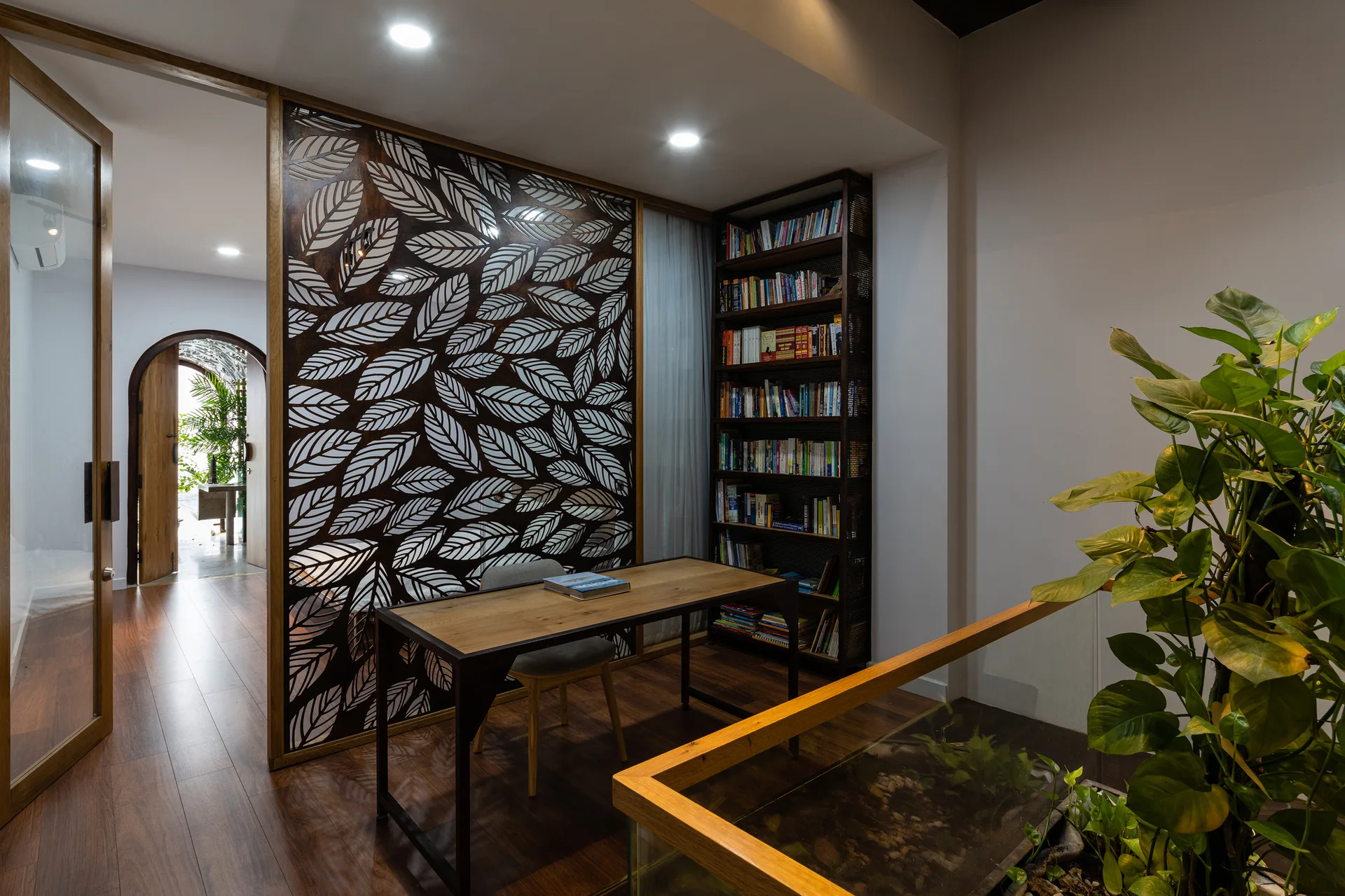 A home office with a wooden desk, bookshelf, and a large decorative screen with a leaf pattern beside a hallway with an arched doorway.