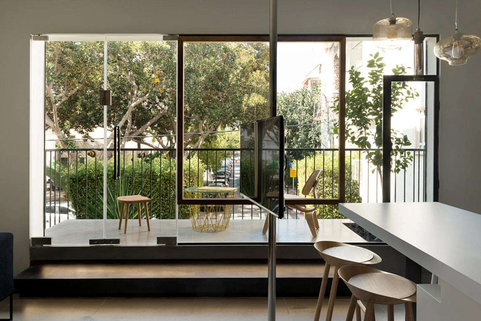 A modern interior features wooden stools at a white counter next to a floor-to-ceiling glass wall that opens onto a balcony with trees.