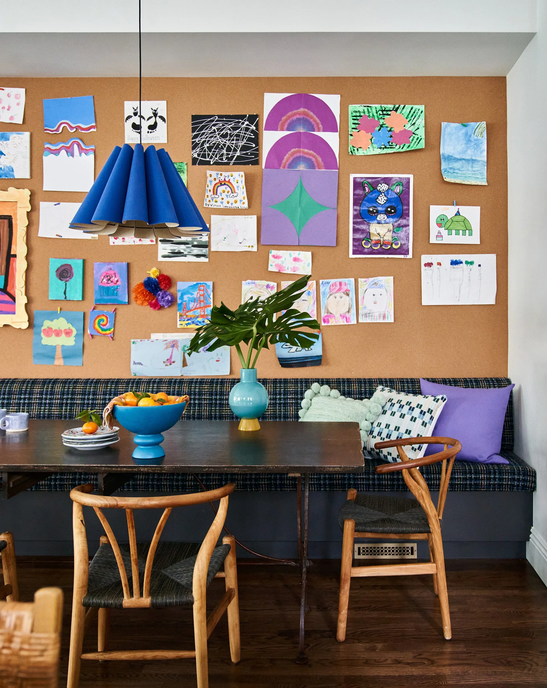 A dining nook features a plaid banquette, wooden wishbone chairs, and a large cork board wall covered in colorful kids' drawings.