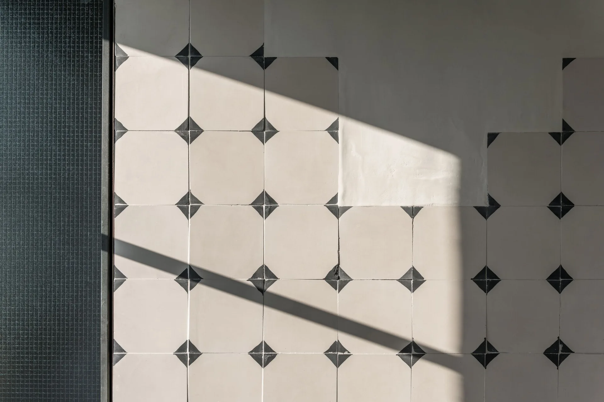 Matte beige square wall tiles feature black geometric corner inserts next to a dark textured glass panel in sharp sunlight.