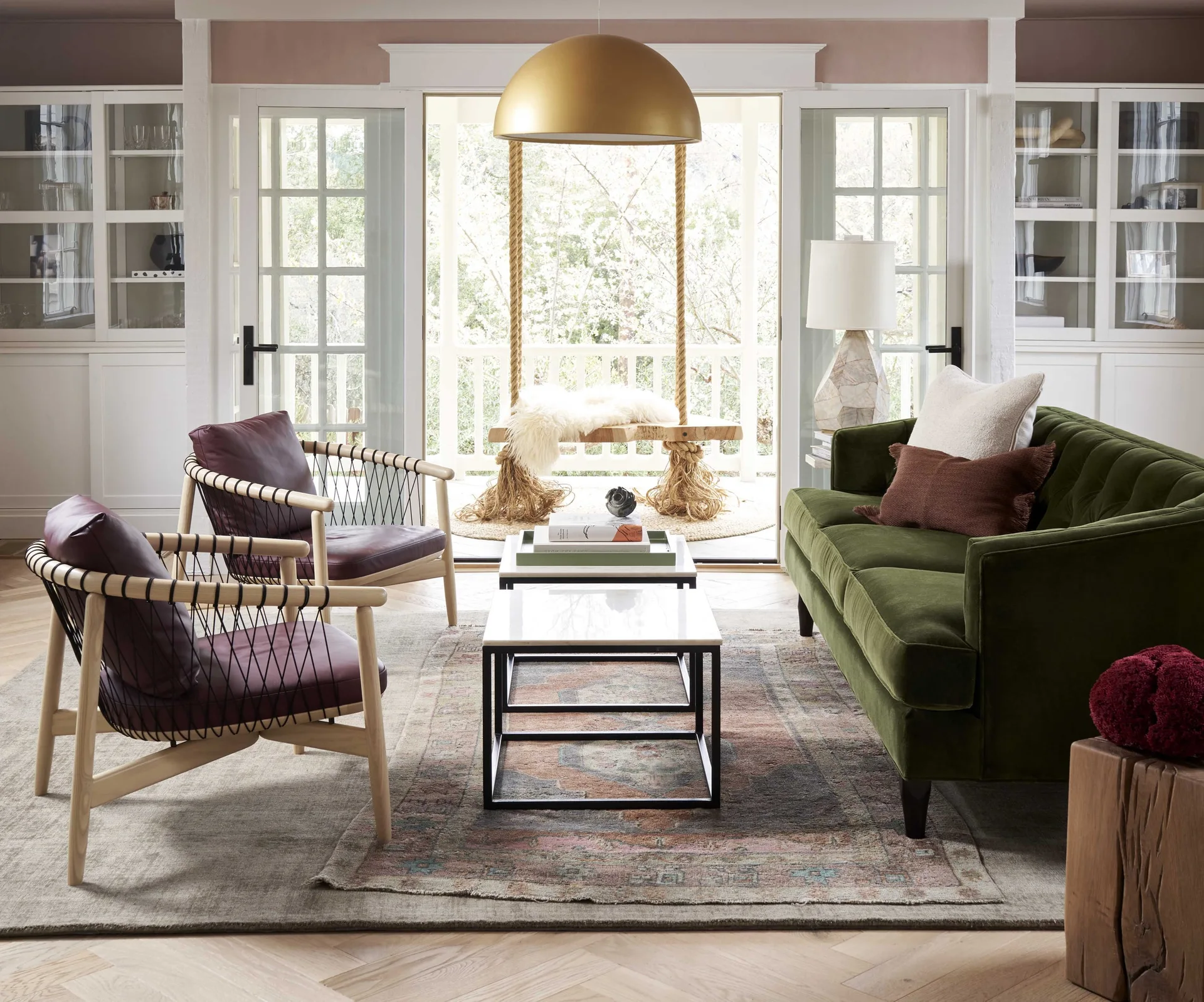 A living room features a green velvet sofa, two leather accent chairs, and nesting coffee tables on a patterned rug in front of French doors.