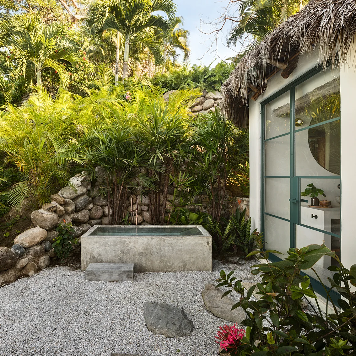 A rectangular concrete soaking tub sits on a gravel path next to a stone retaining wall and lush tropical plants.