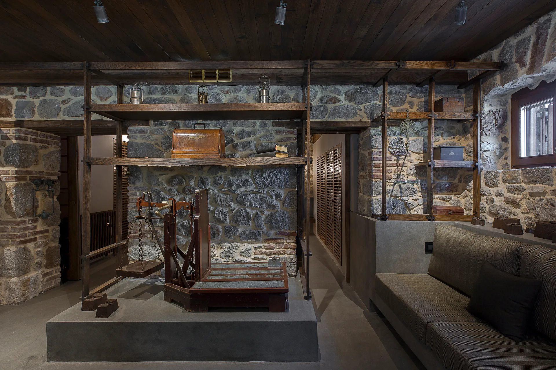 A rustic room featuring exposed stone walls, dark wood plank ceilings, open wooden display shelving, and an antique balance scale on a concrete platform.