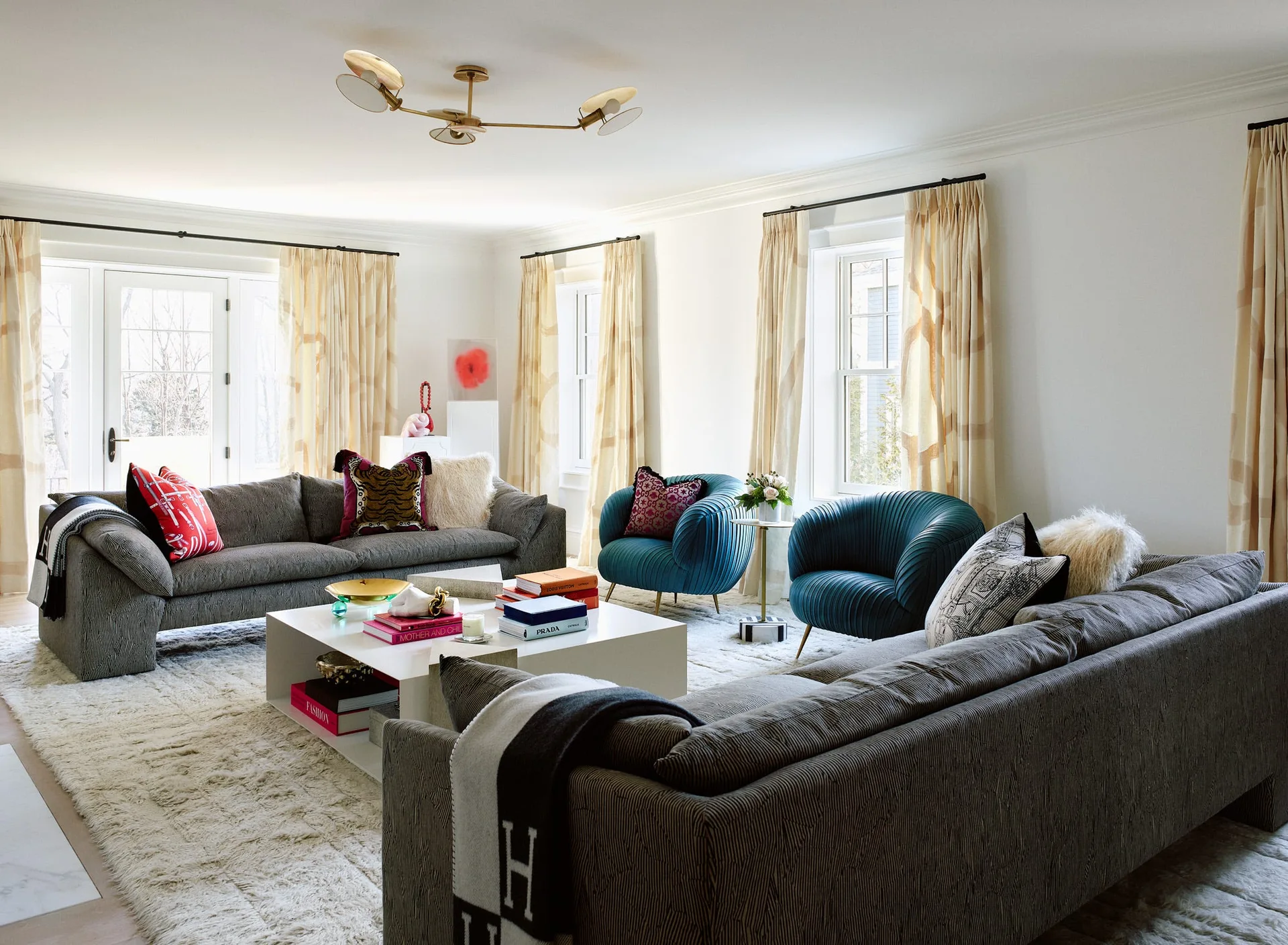 A spacious white living room features two large gray sofas facing each other on a plush rug next to two curved teal armchairs.