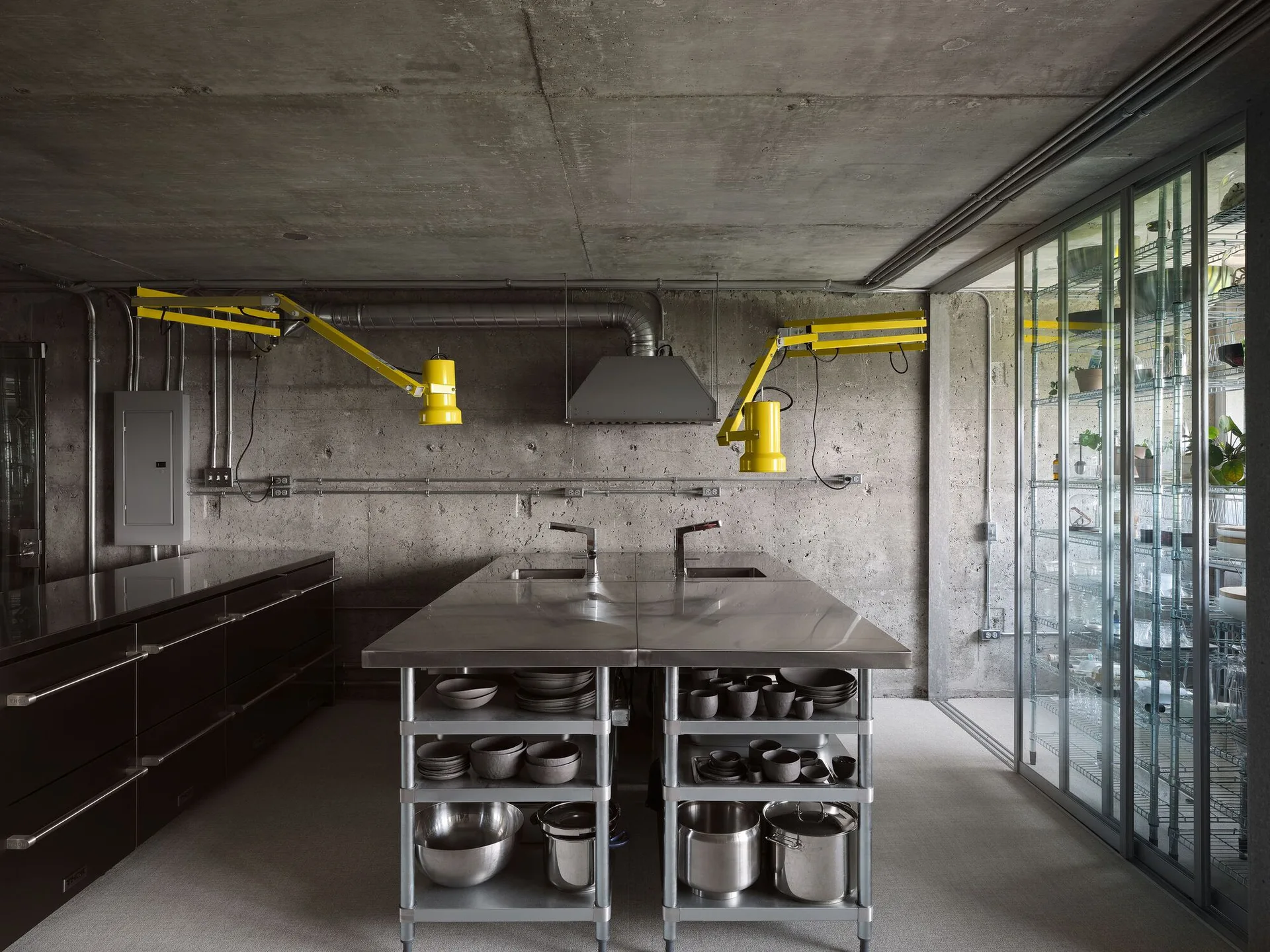 An industrial kitchen featuring a stainless steel island, raw concrete walls and ceiling, dark cabinets, and two yellow adjustable swing-arm lamps.