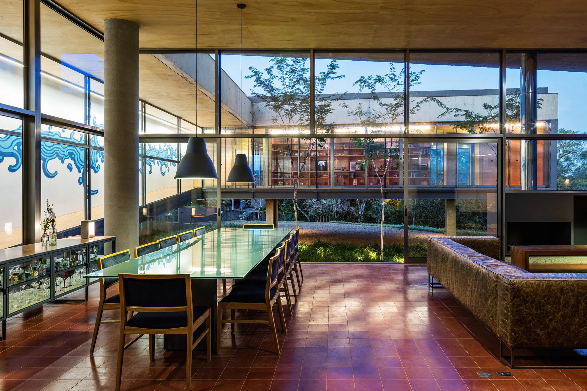 A spacious modern dining room features a long glass-top table, wooden chairs, a leather sofa, and floor-to-ceiling windows looking out to a courtyard.
