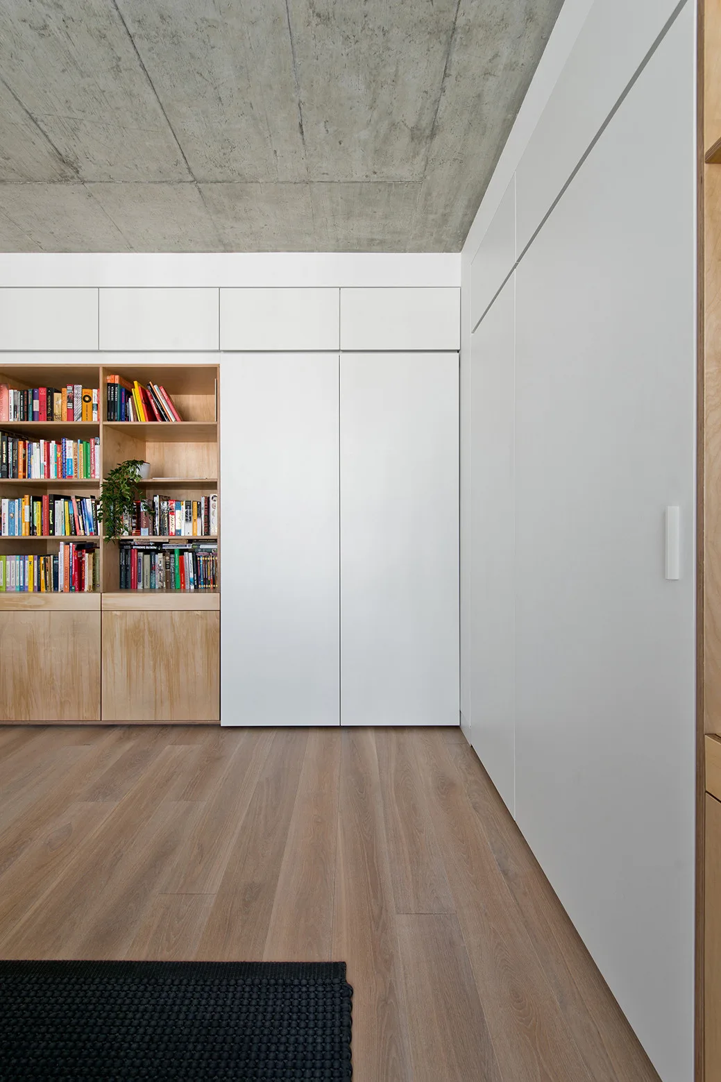 A modern room featuring floor-to-ceiling white storage cabinets integrated with a light wood bookshelf under an exposed concrete ceiling.
