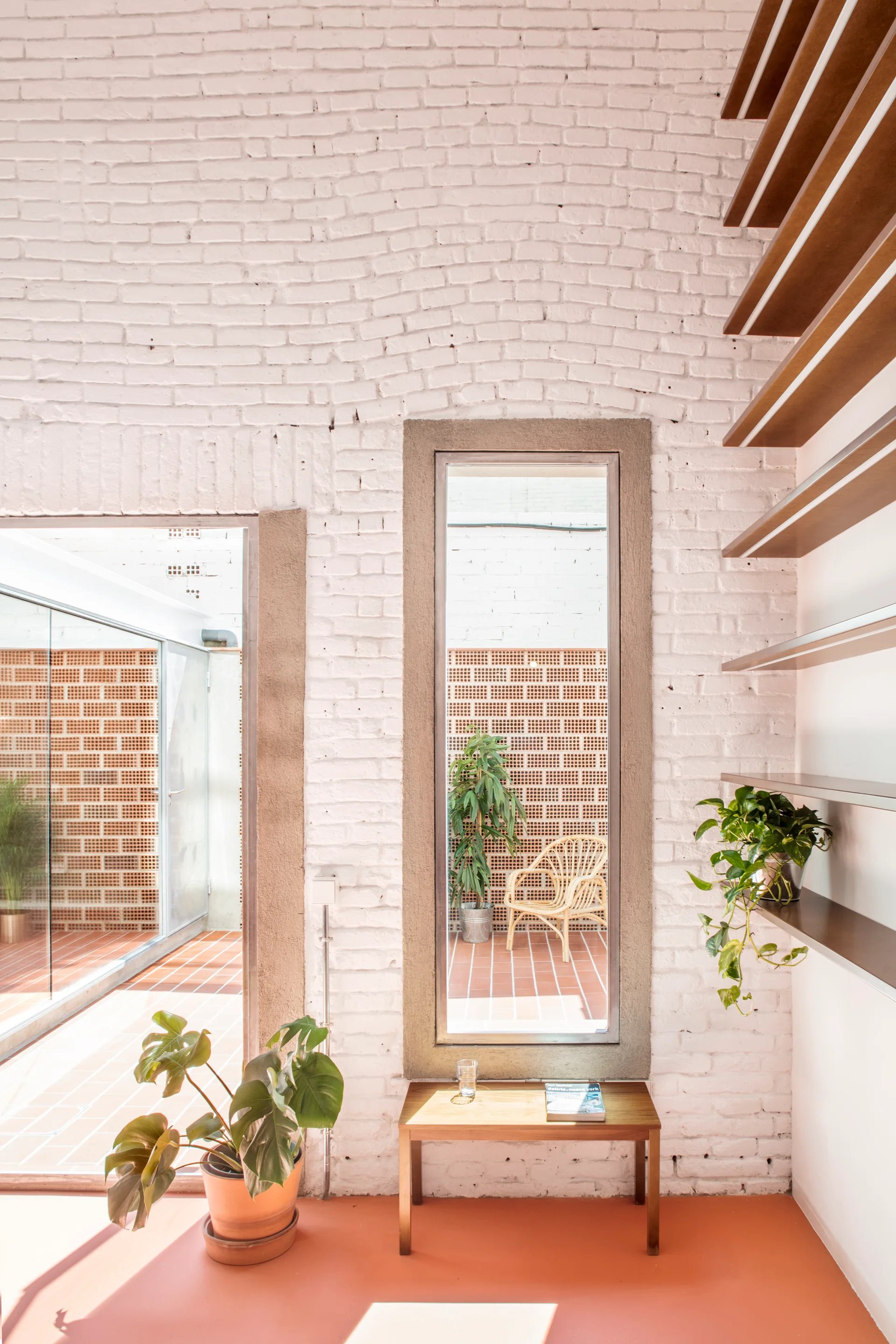 An interior space with painted white brick walls, terracotta floors, wooden shelves, and a narrow window looking out to a courtyard.
