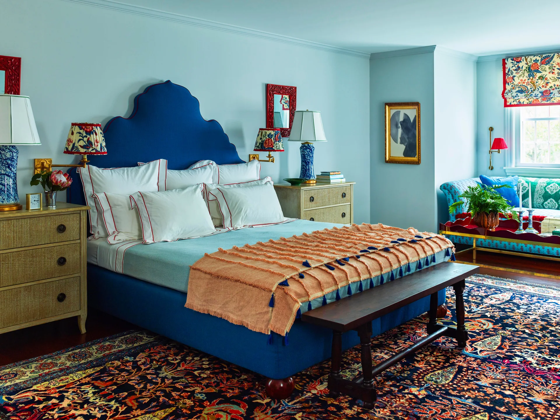 An eclectic bedroom features a blue scalloped headboard bed, textured rattan nightstands, an orange fringed throw, and a colorful patterned area rug.