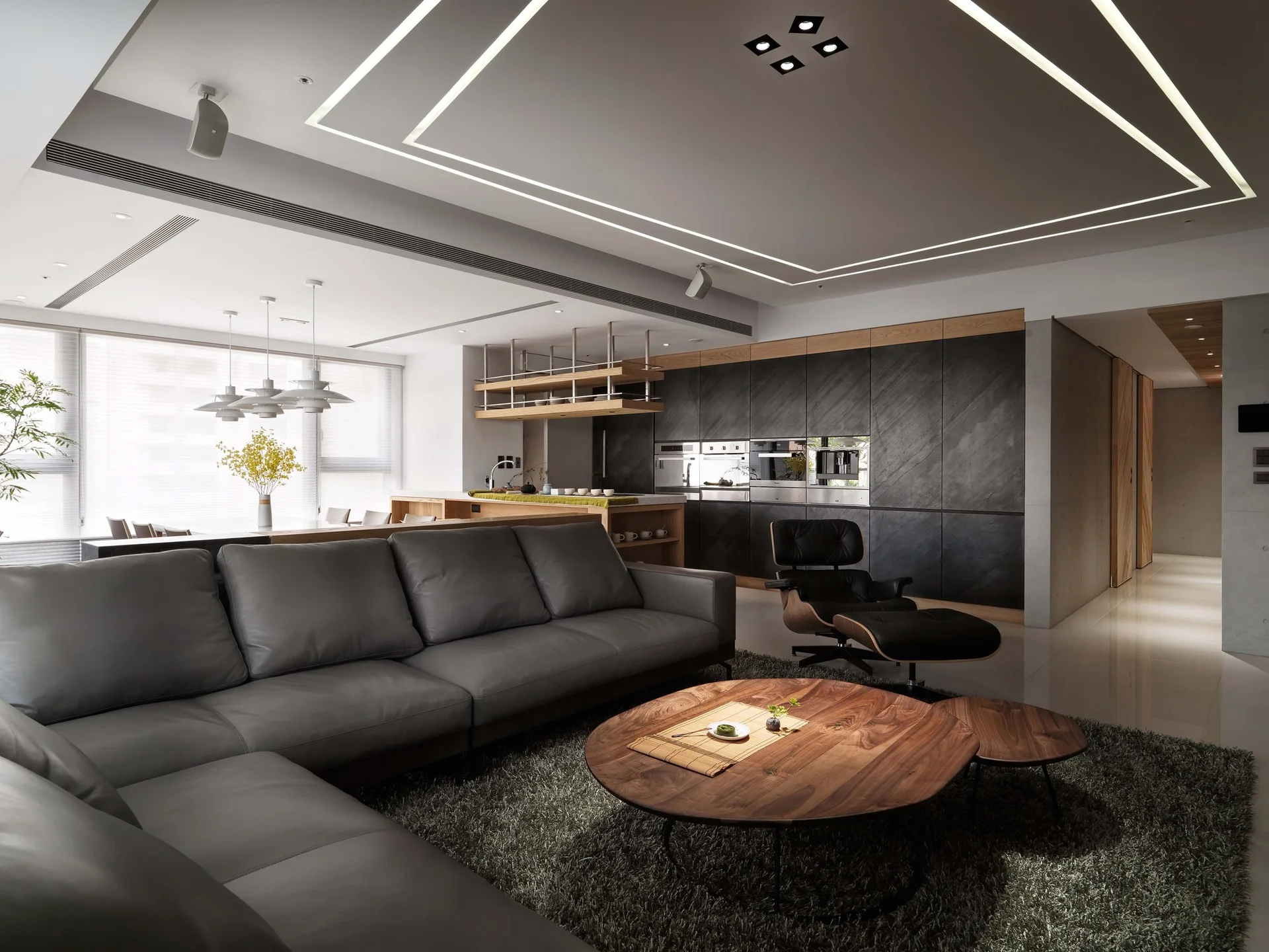 A contemporary open-plan living room features a grey leather sectional sofa, a round wood coffee table, and a dark grey kitchen wall in the background.