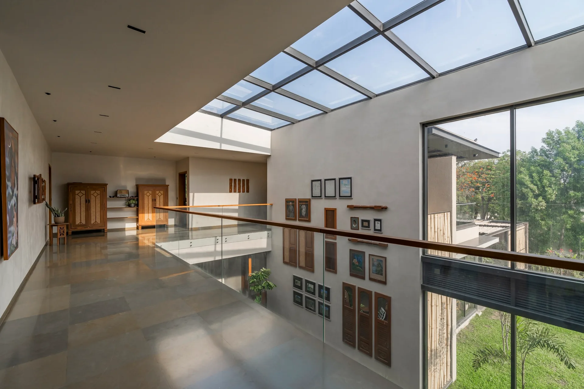 A spacious interior landing features polished stone floors, a glass-paneled railing with a wood cap, a grid skylight, and a double-height gallery wall.