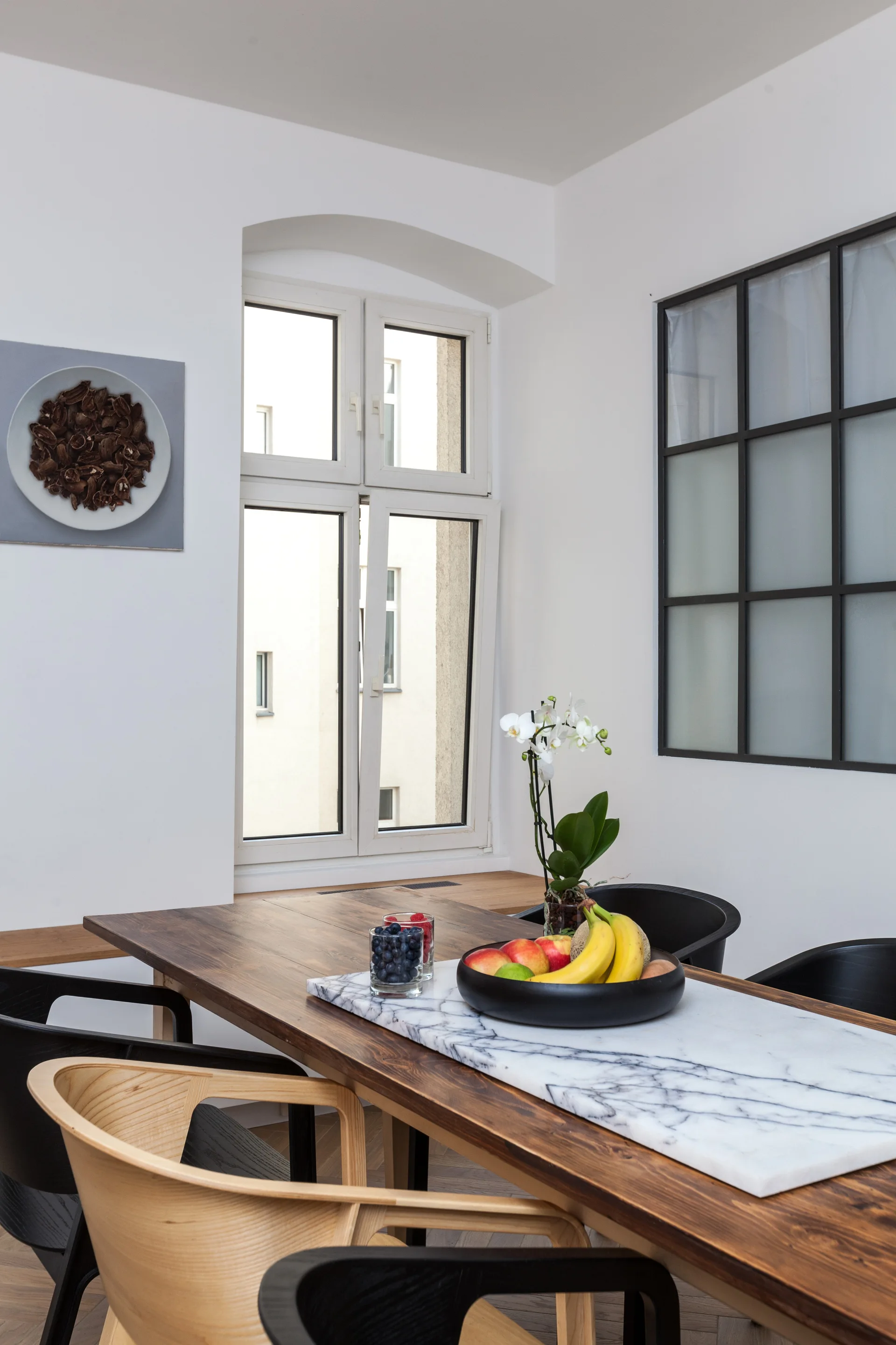 A wooden dining table featuring a white marble inlay is surrounded by black and light wood chairs in a bright minimalist dining room.